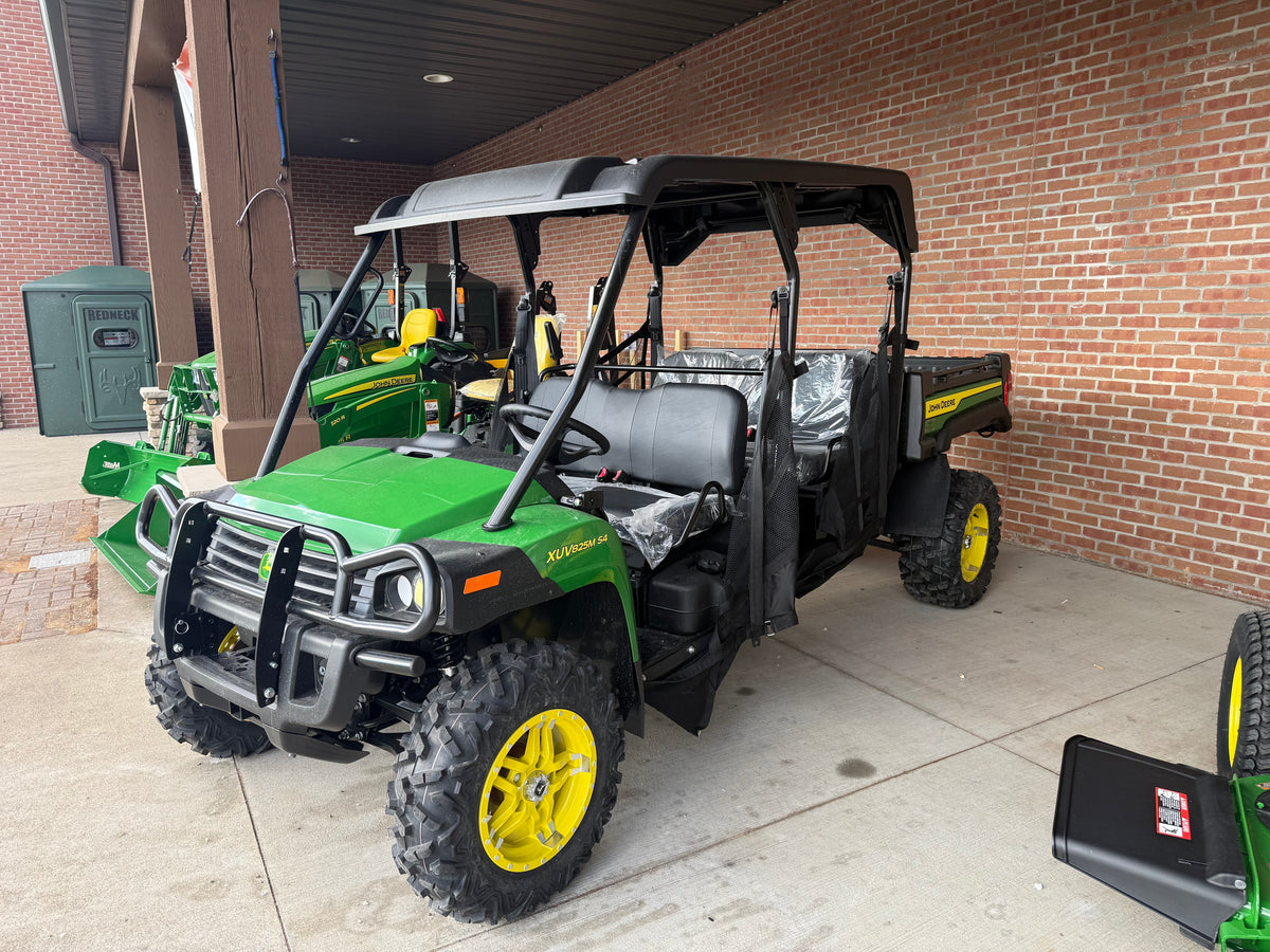 Green and yellow John Deere four-seater utility vehicle with off-road tires and protective canopy, parked on concrete near a brick wall at a dealership.