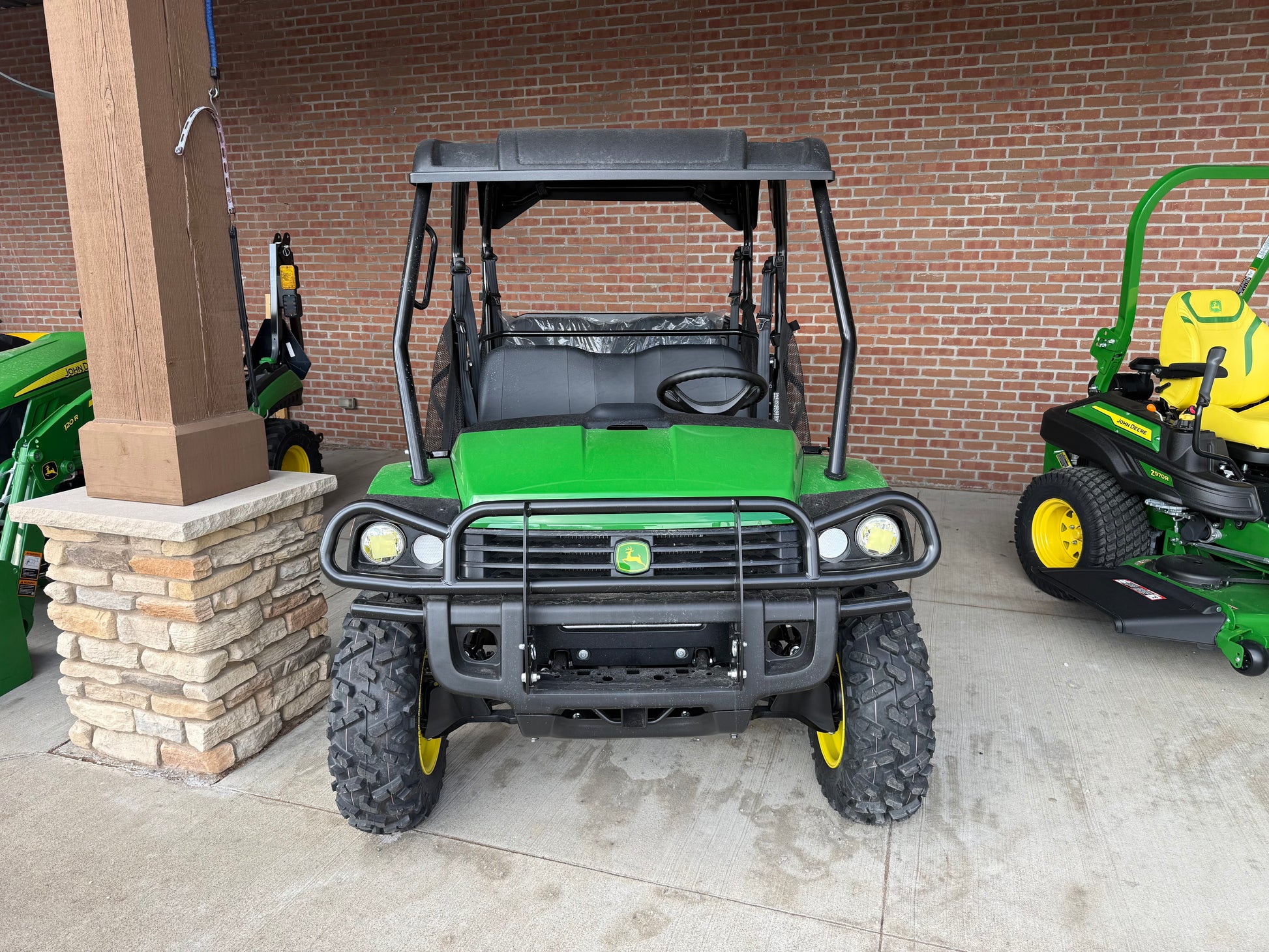 Front view of a green John Deere 4-seater crossover utility vehicle with rugged tires and protective roof, displayed outdoors near other equipment.