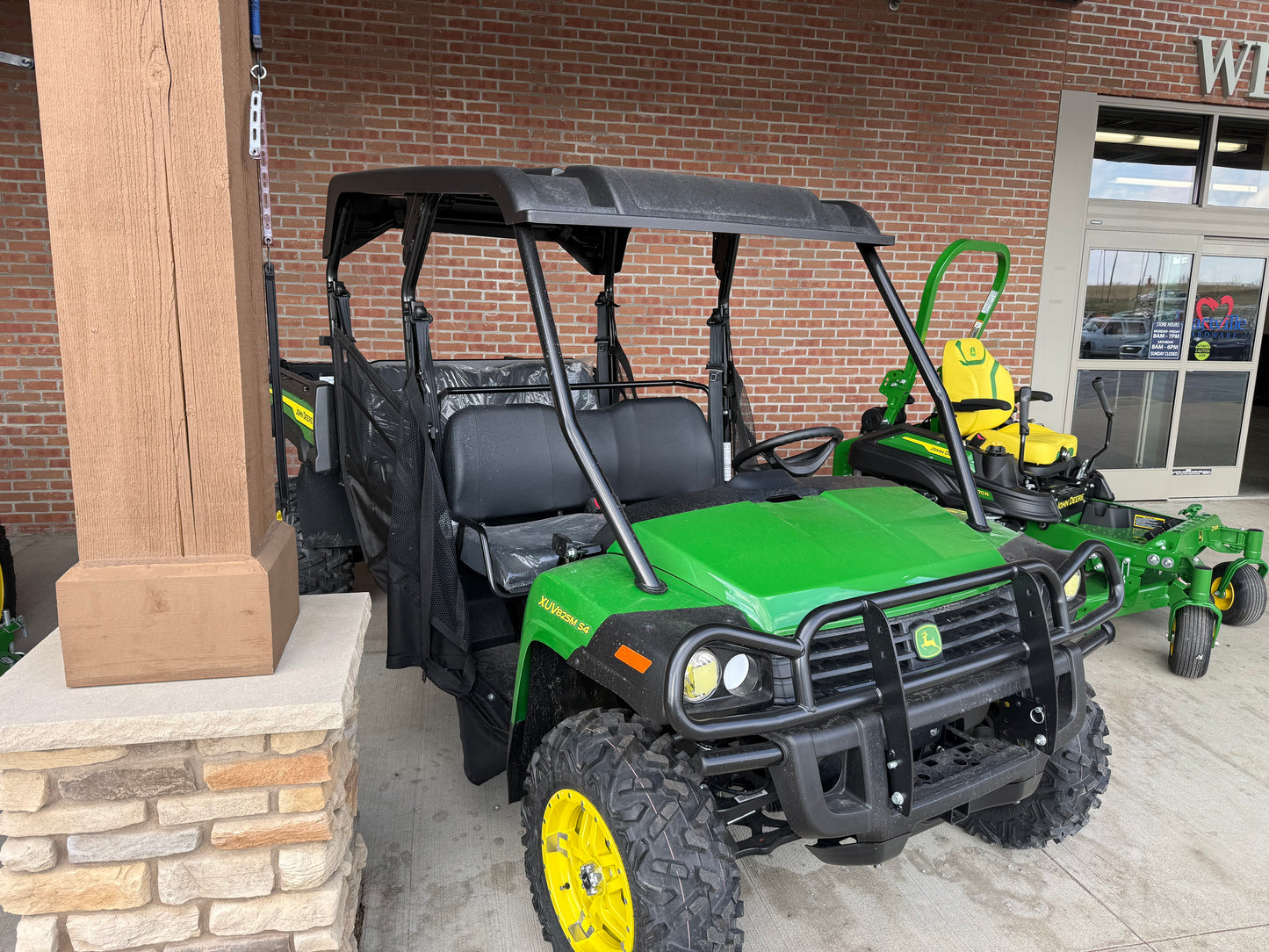 Green John Deere four-seater crossover utility vehicle with rugged tires and protective canopy, displayed outdoors at a dealership among other equipment.
