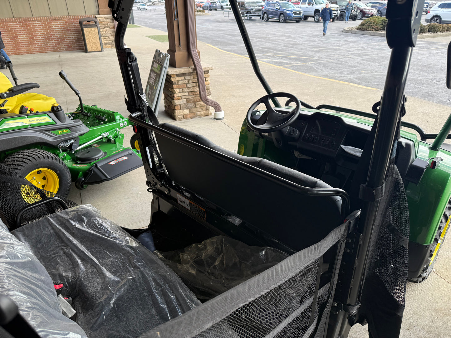 Rear interior view of a green John Deere 4-seater utility vehicle showing bench seating with protective covers and dashboard in a dealership lot among equipment
