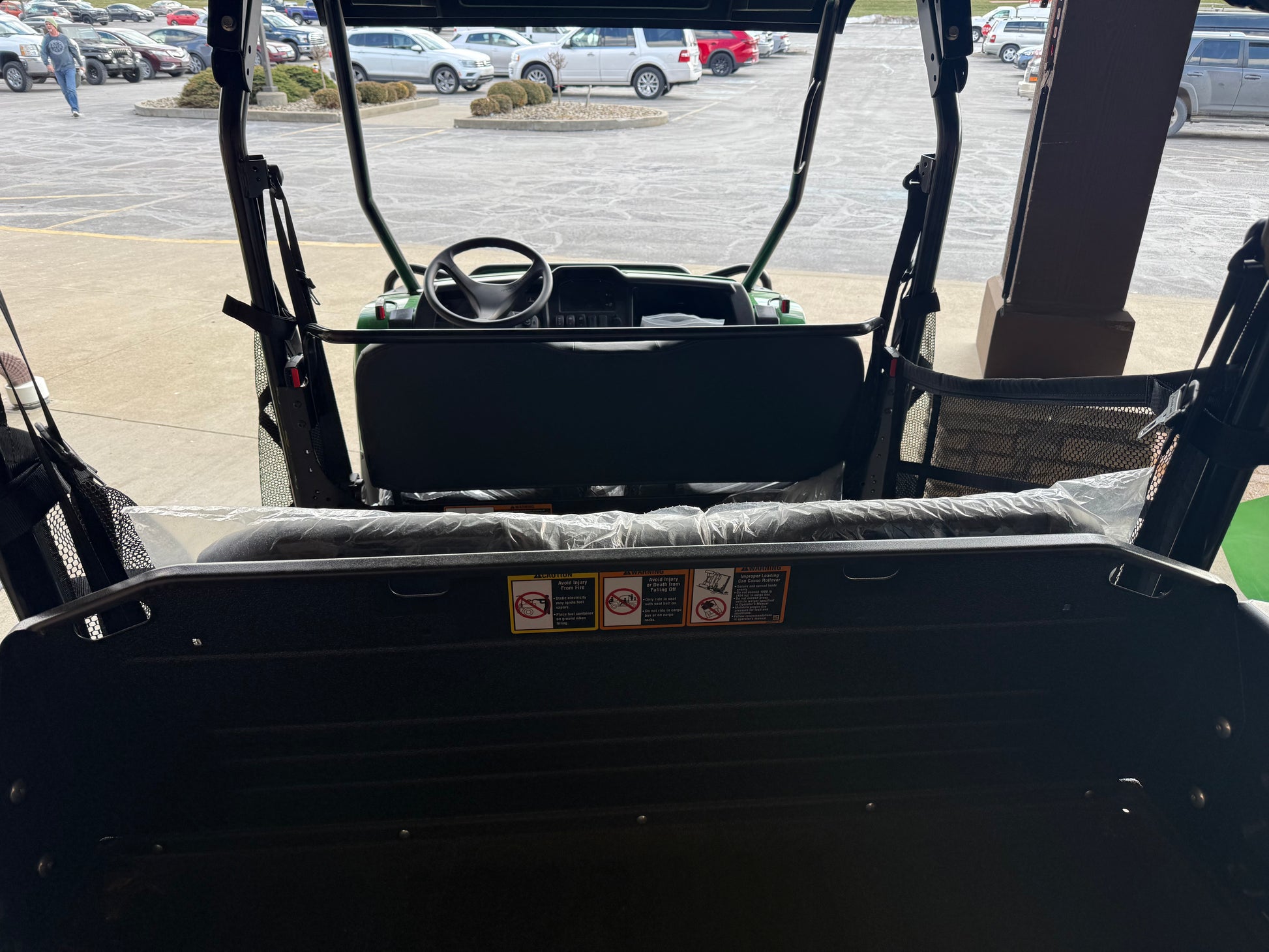 View from the rear bed area of a John Deere four-seater crossover utility vehicle showing steering wheel, dashboard, and protective covers on seats in a dealership lot.