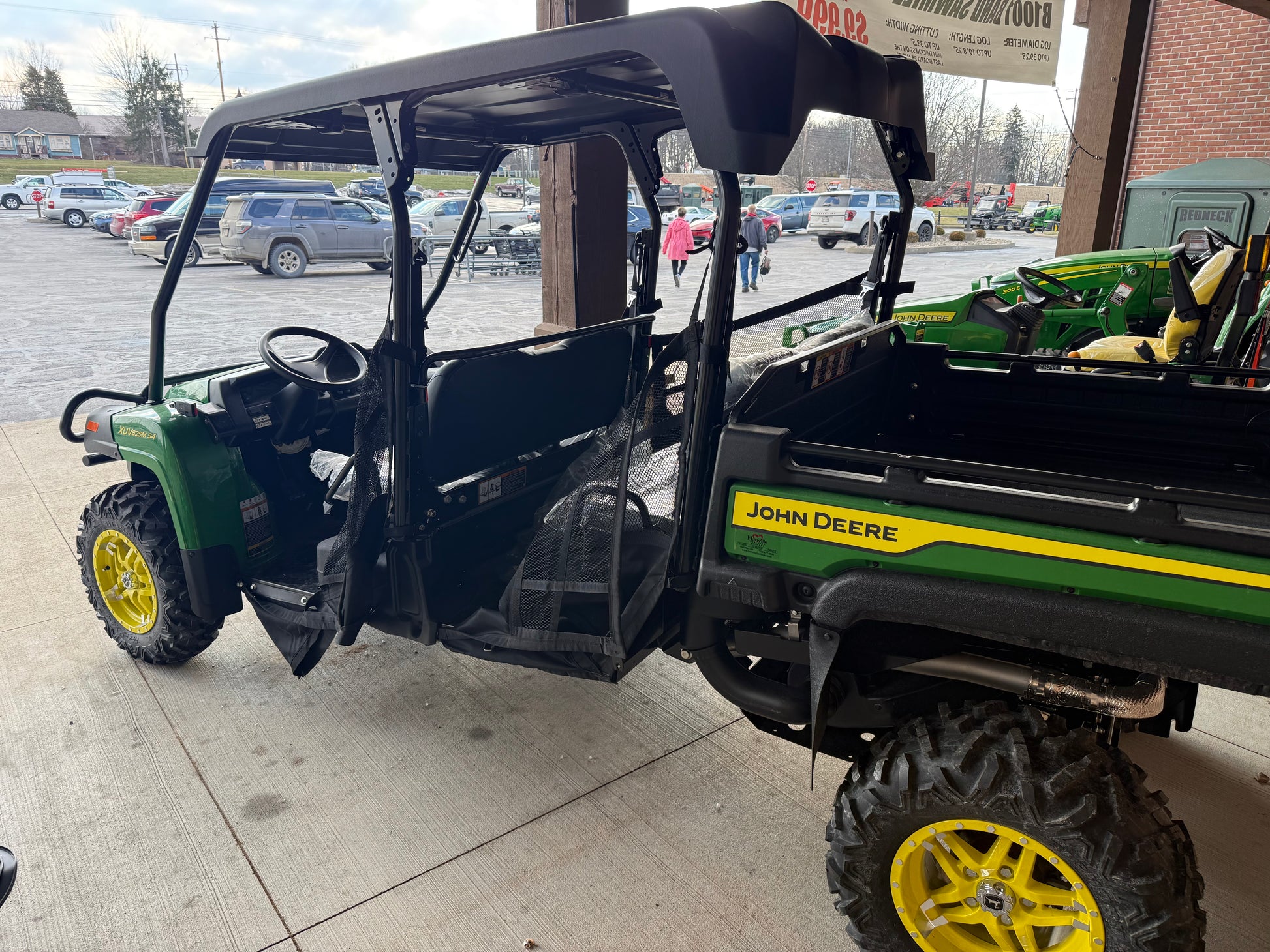 John Deere green and yellow 4-seater crossover utility vehicle with rugged tires and open cabin, parked near equipment at a dealership lot.