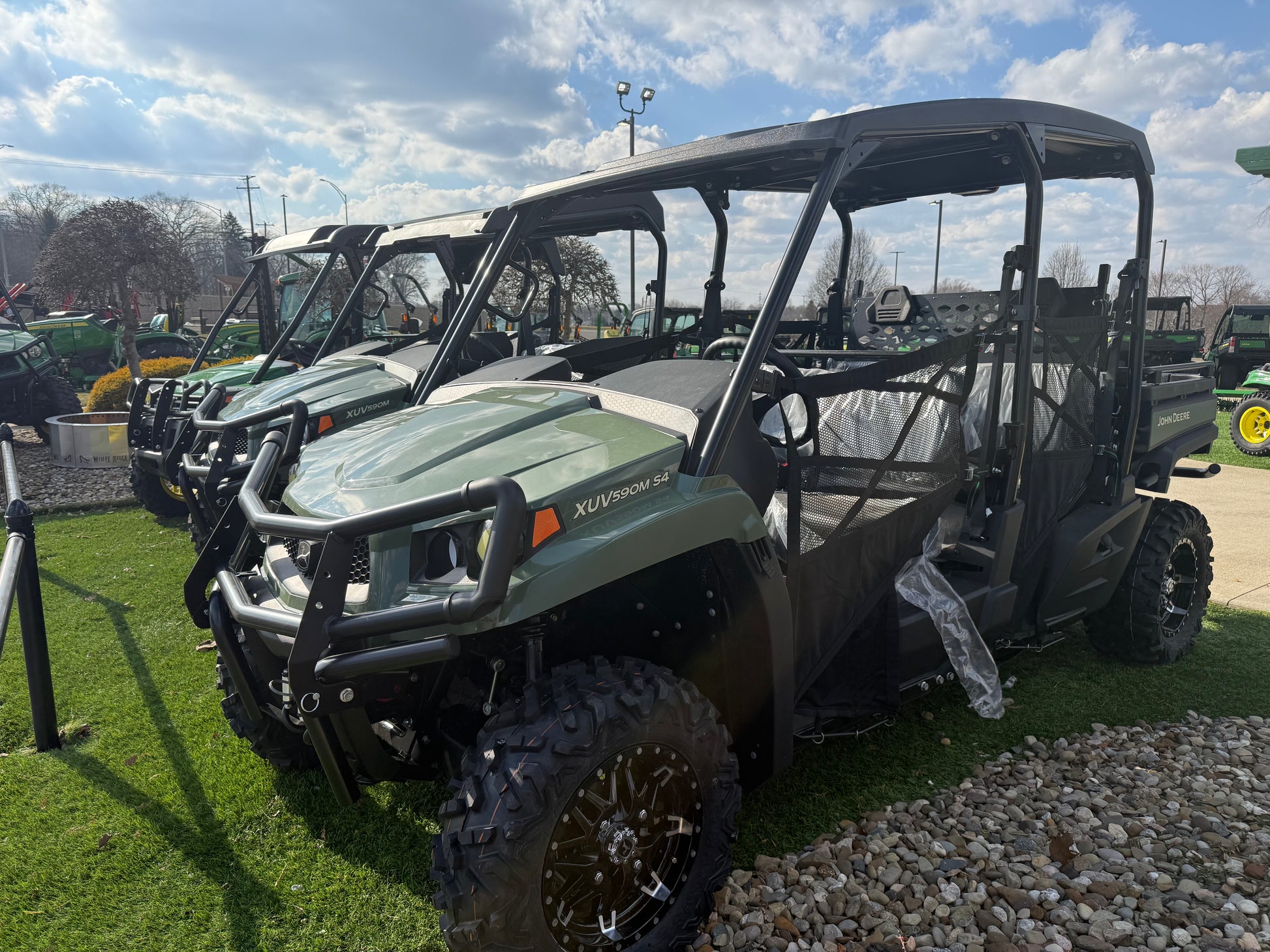 Four-seater crossover utility vehicle with rugged tires and protective canopy, parked outdoors on grass and gravel near other similar vehicles at a dealership