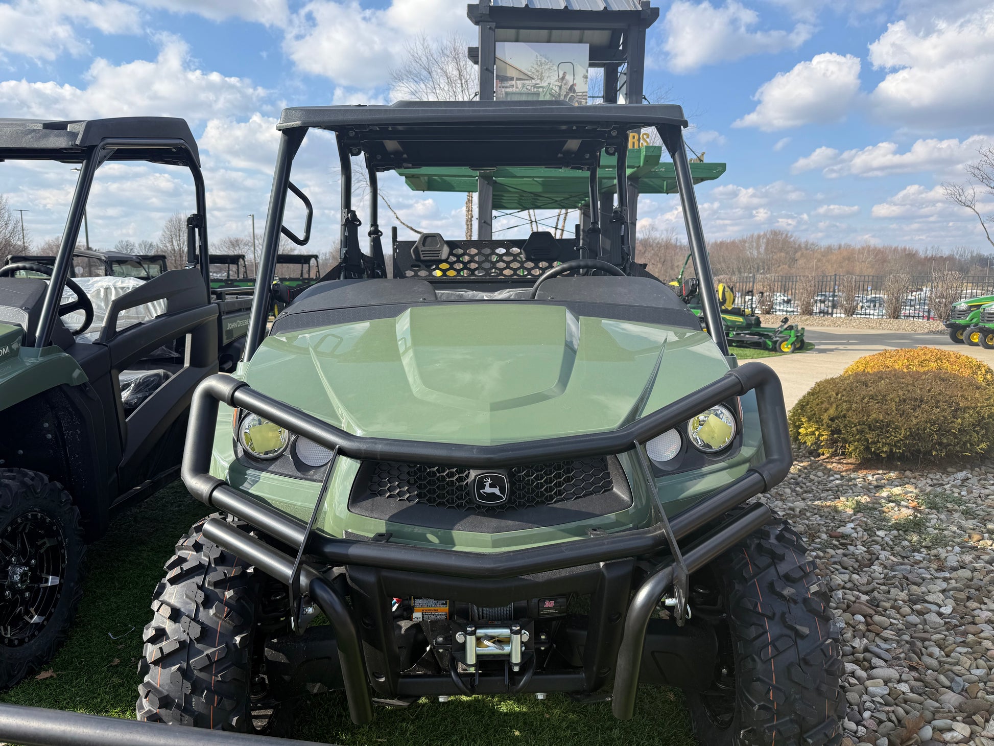 Front view of a green John Deere 4-seater crossover utility vehicle with rugged tires and reinforced bumper, displayed outdoors among other UTVs and equipment