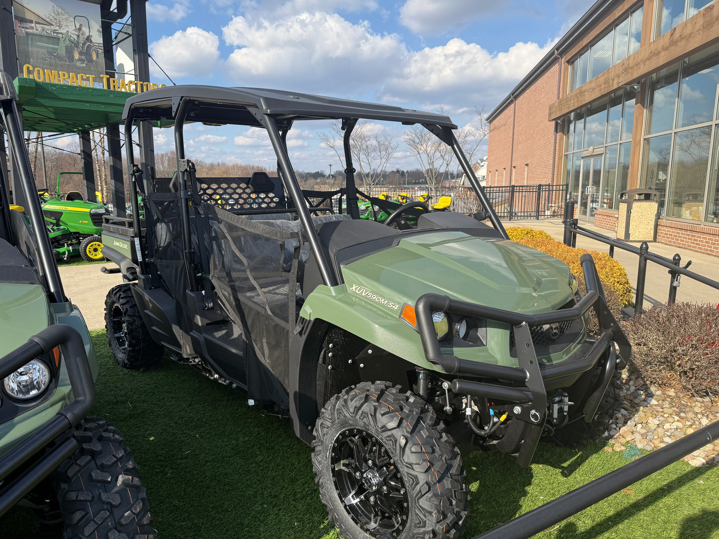 Green John Deere four-passenger crossover utility vehicle with rugged tires and reinforced front bumper, displayed outdoors near dealership building and other equipment.