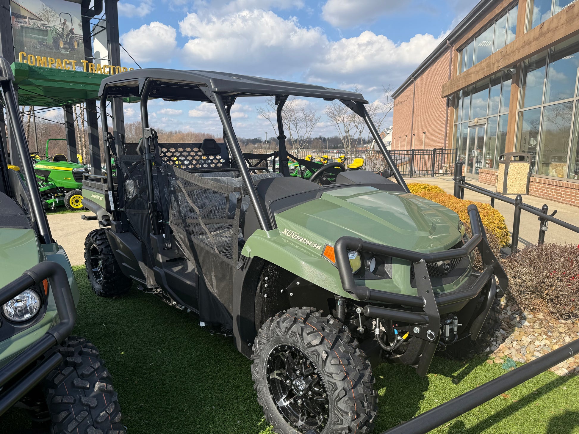 Green John Deere four-passenger crossover utility vehicle with rugged tires and reinforced front bumper, displayed outdoors near dealership building and other equipment.