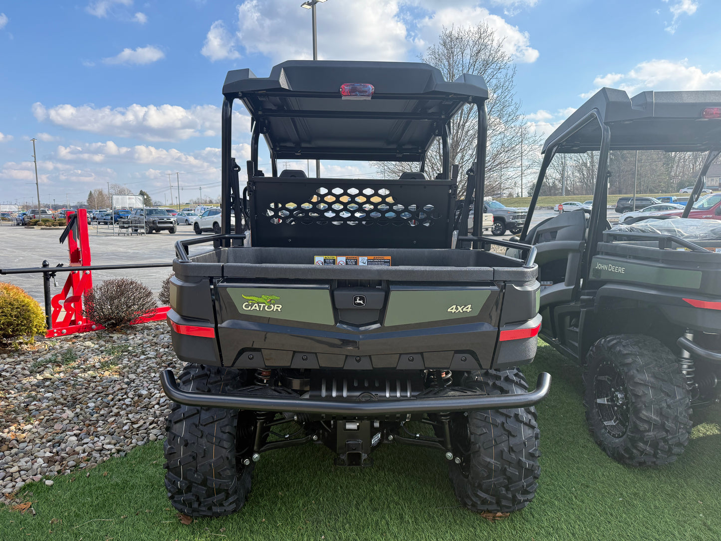 Rear view of a green and black John Deere 4-seater crossover utility vehicle with rugged tires and protective roof, parked on artificial grass near other equipment.