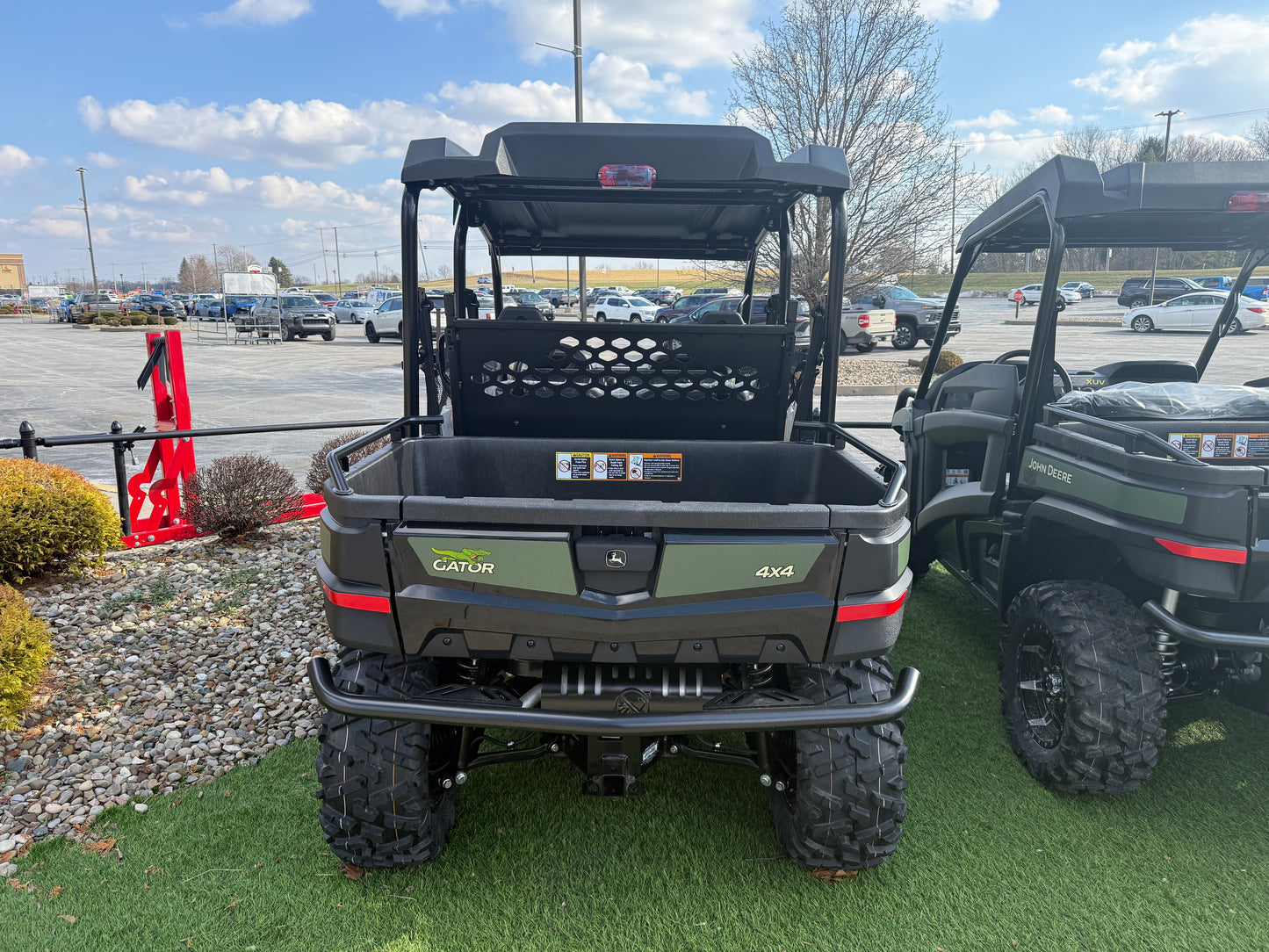 Rear view of a John Deere 4x4 Gator utility vehicle with cargo bed, rugged tires, and protective roof, displayed outdoors near other equipment.