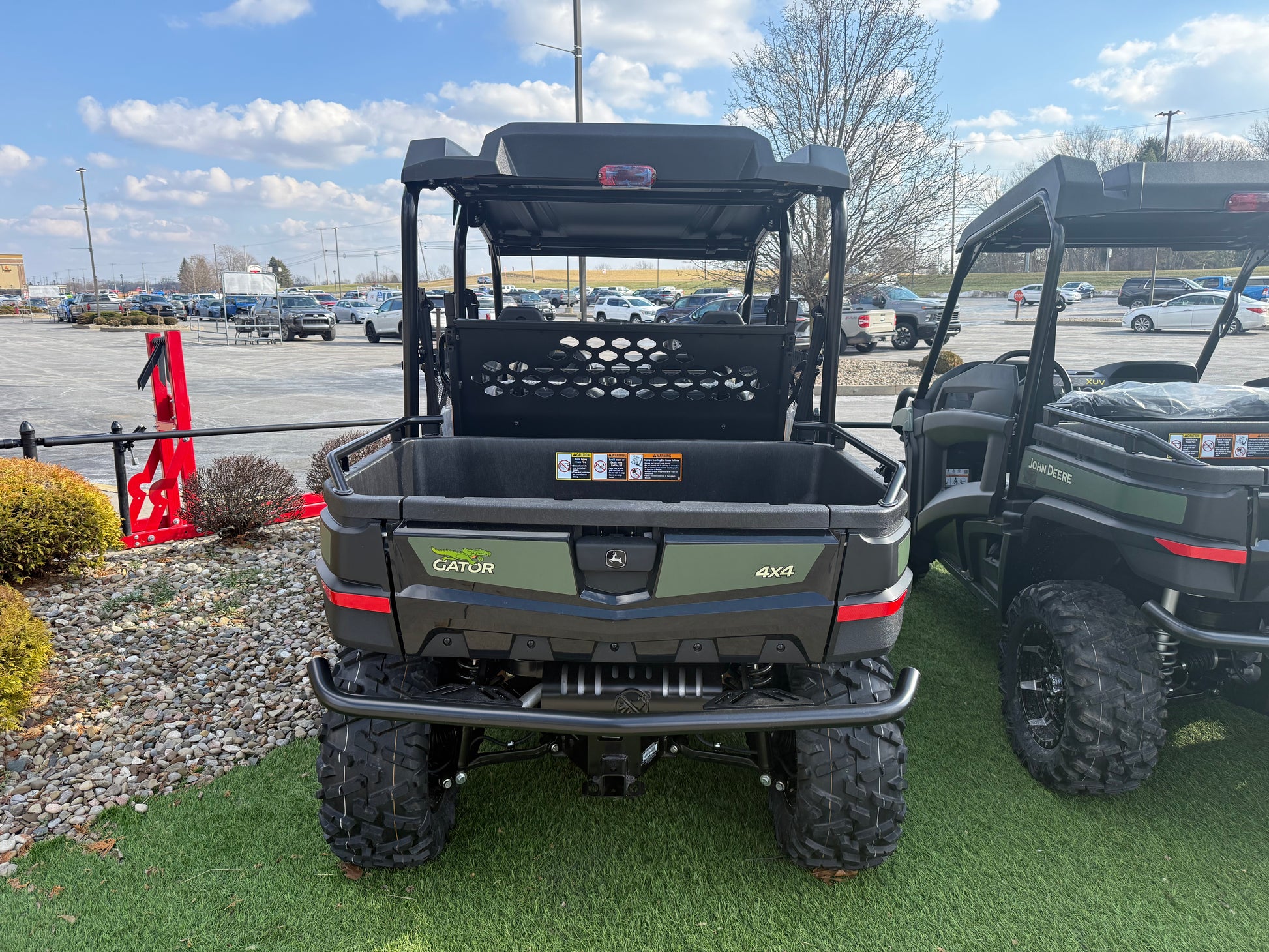 Rear view of a John Deere 4x4 Gator utility vehicle with cargo bed, rugged tires, and protective roof, displayed outdoors near other equipment.