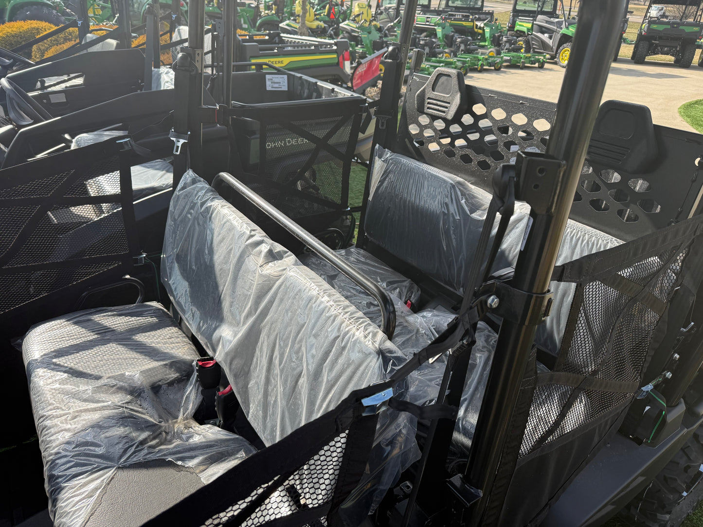 Interior view of a John Deere four-passenger crossover utility vehicle featuring bench seats protected with plastic covers and safety netting in a dealership lot.
