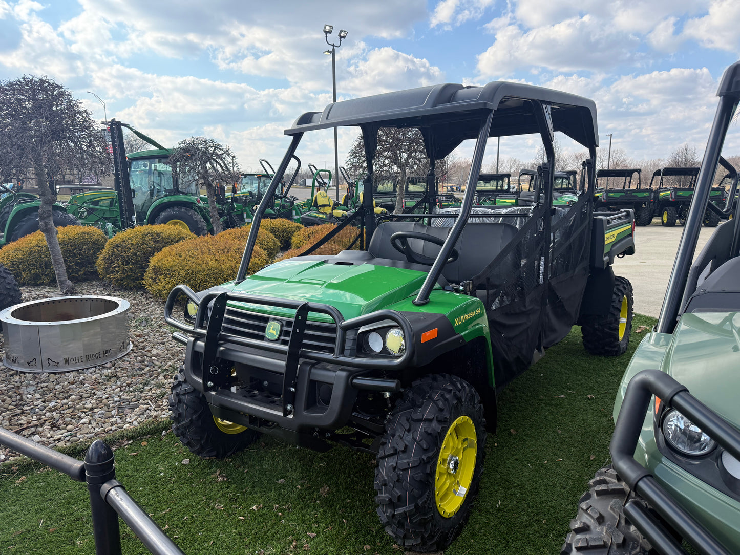 Green and black John Deere 4-seater crossover utility vehicle with rugged tires and protective roof parked outdoors on grass near other equipment at dealership