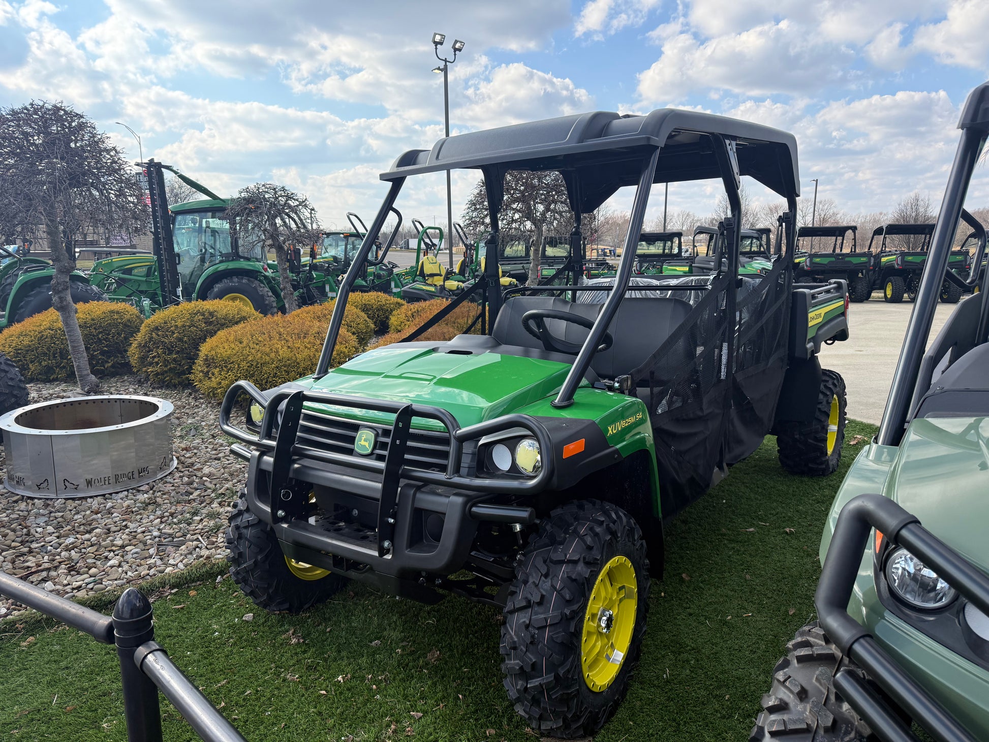 Green and black John Deere 4-seater crossover utility vehicle with rugged tires and protective roof parked outdoors on grass near other equipment at dealership