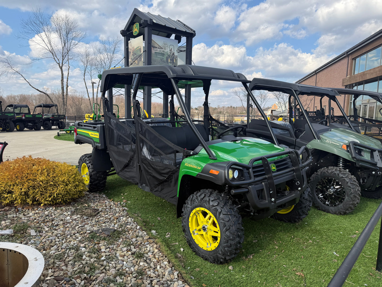 Green John Deere 4-seater crossover utility vehicle with protective soft doors and rugged tires displayed outdoors on artificial grass near other UTVs at dealership