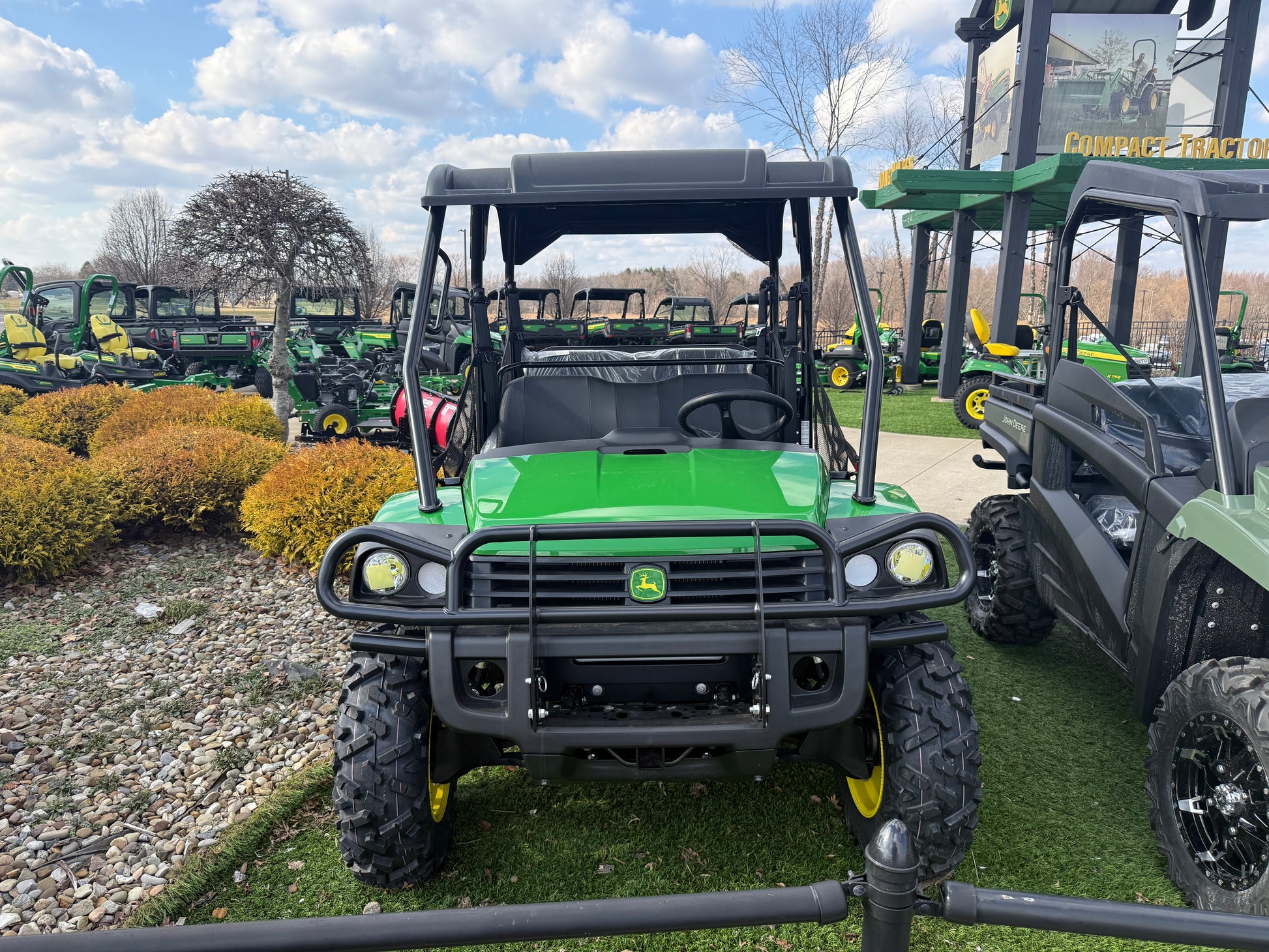 Front view of a green John Deere 4-seater crossover utility vehicle with protective roof and rugged tires, displayed outdoors among other John Deere Equipment.