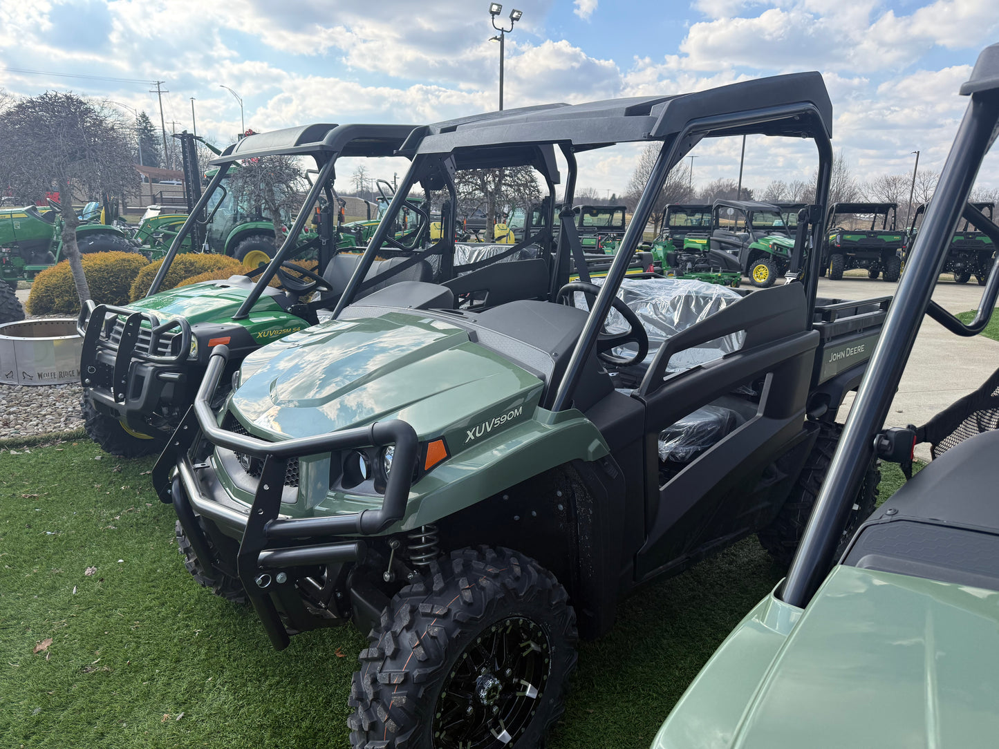 Green John Deere mid-size crossover utility vehicle with protective roof and rugged tires displayed outdoors on artificial grass at dealership lot.