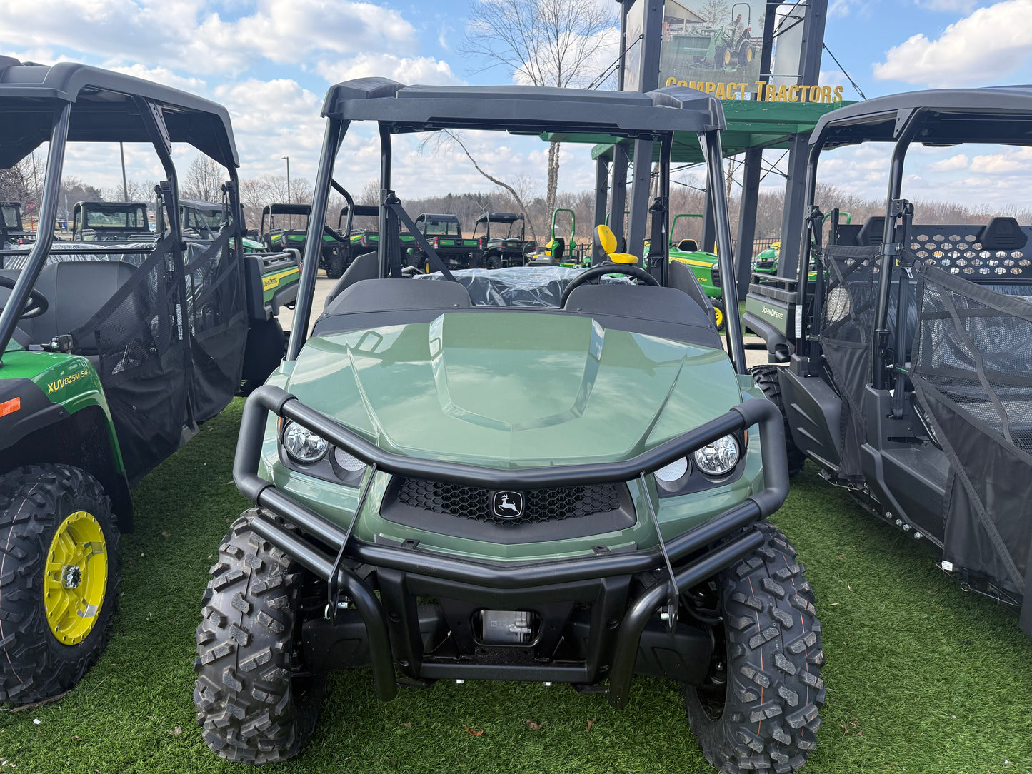 Front view of a green mid-size John Deere utility vehicle with rugged tires and protective roof, displayed outdoors on artificial grass alongside other UTVs.