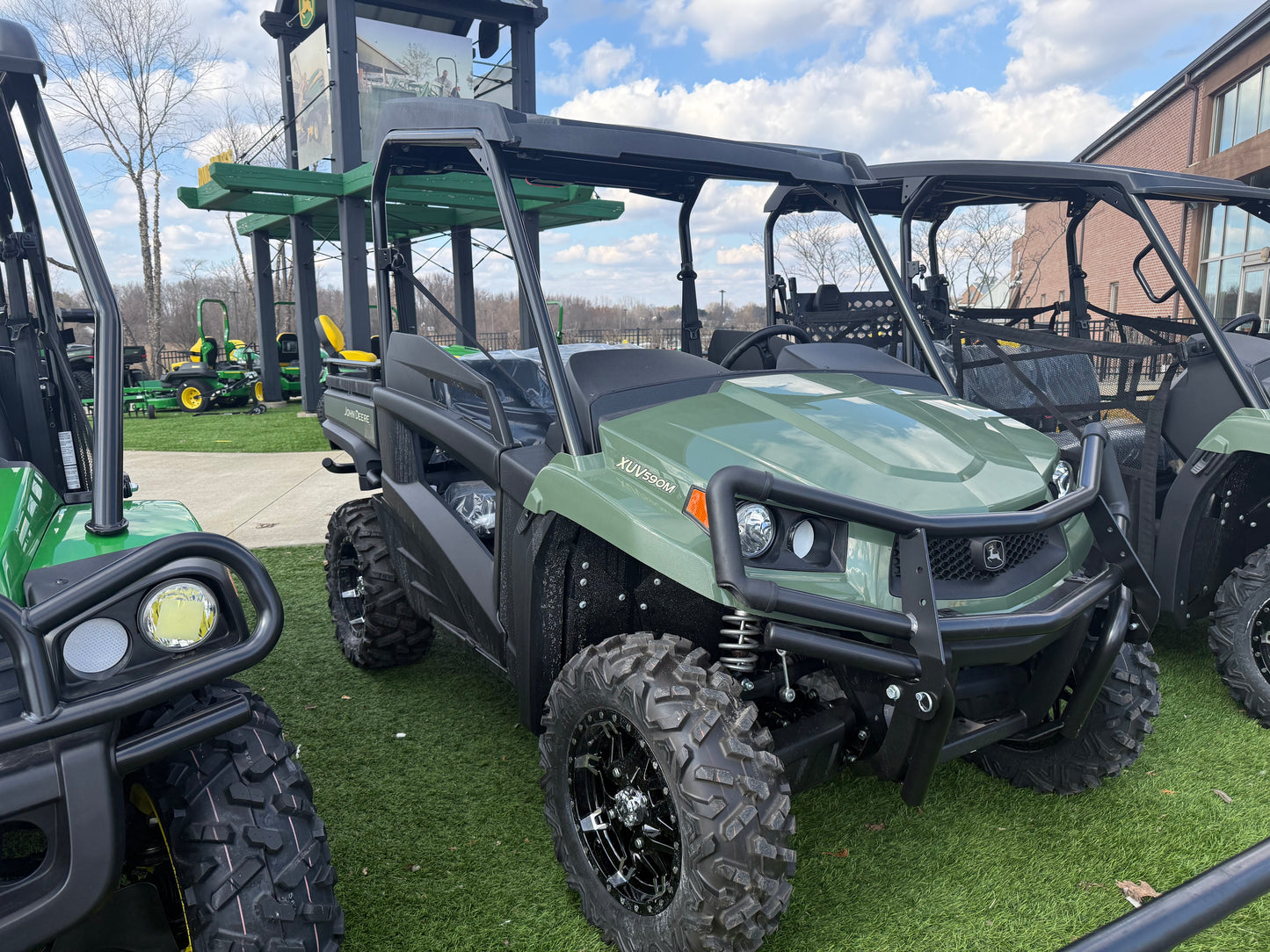 Green mid-size John Deere crossover utility vehicle with protective roll cage and aggressive off-road tires displayed outdoors on artificial grass at dealership lot.