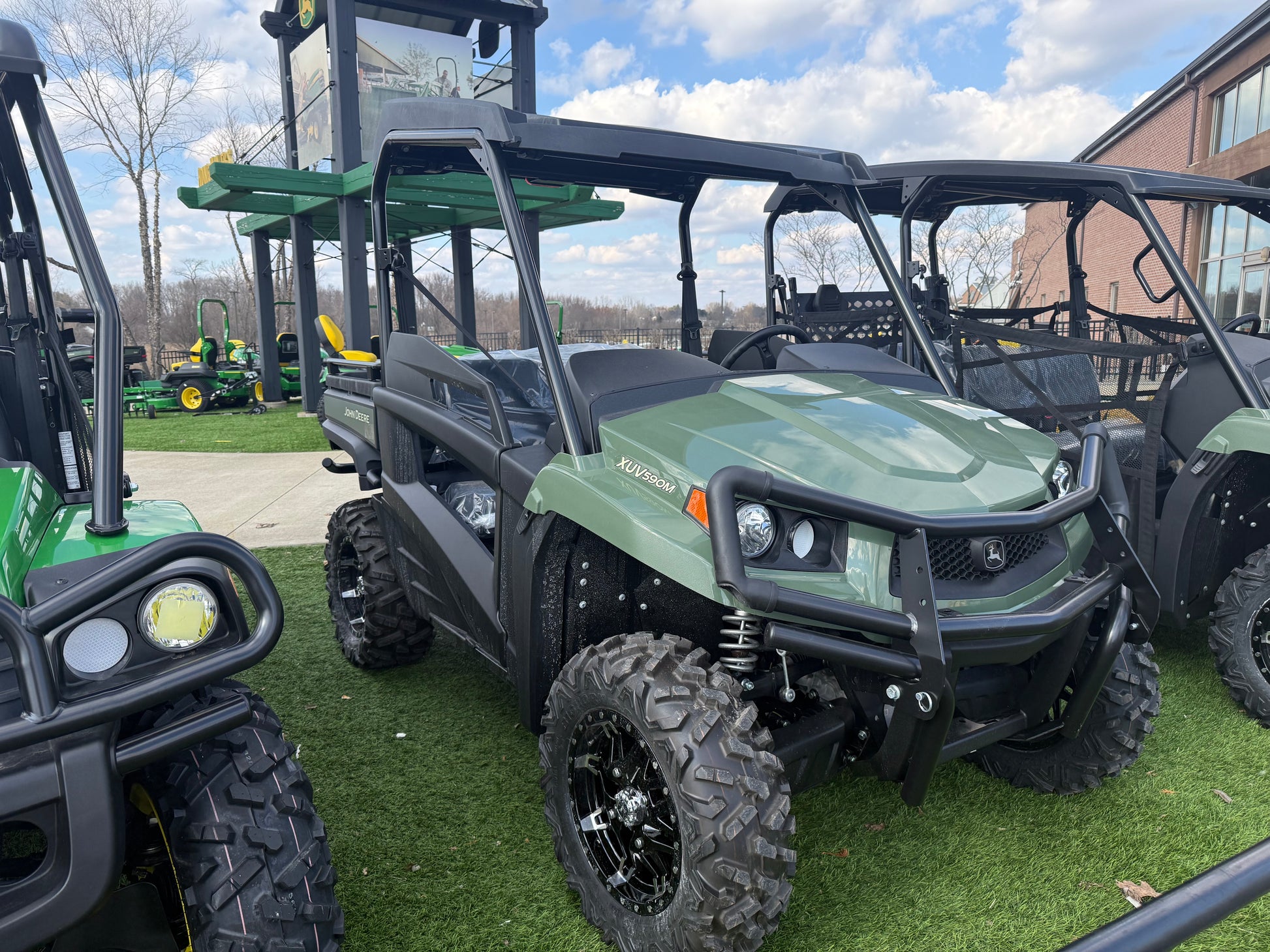 Green mid-size John Deere crossover utility vehicle with protective roll cage and aggressive off-road tires displayed outdoors on artificial grass at dealership lot.