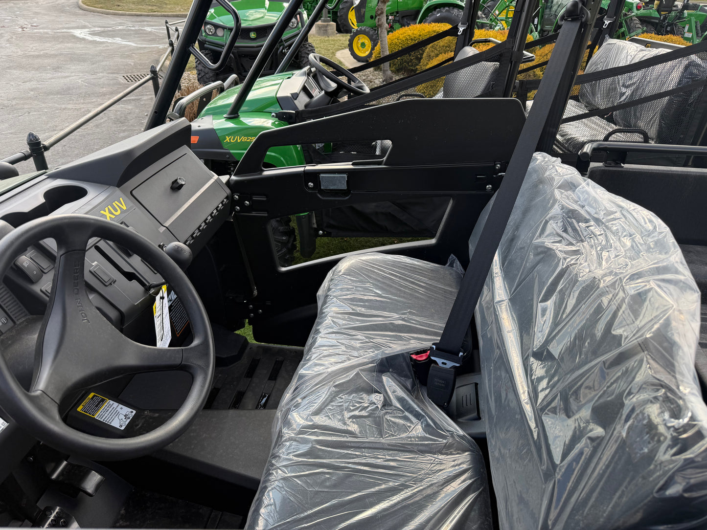 Interior view of a mid-size John Deere crossover utility vehicle showing the steering wheel, dashboard, and seats covered in protective plastic at a dealership lot.