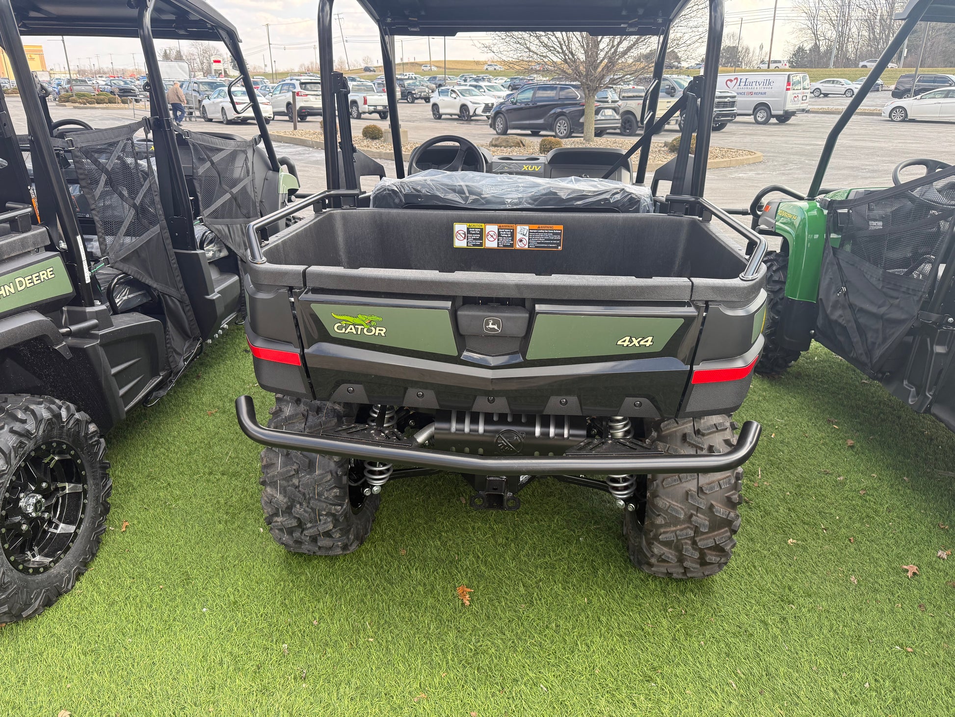 Rear view of a John Deere 4x4 utility vehicle with a cargo bed, rugged off-road tires, and protective bumper, displayed on artificial grass at dealership lot