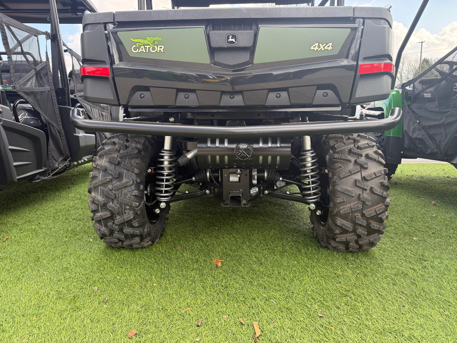 Rear close-up of a green John Deere Gator 4x4 utility vehicle showing suspension, rugged tires, and protective rear bumper on artificial grass at dealership lot