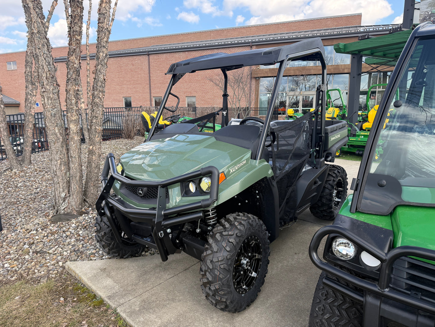 Green mid-size crossover utility vehicle with off-road tires and protective roll cage parked on concrete near dealership building and other equipment.