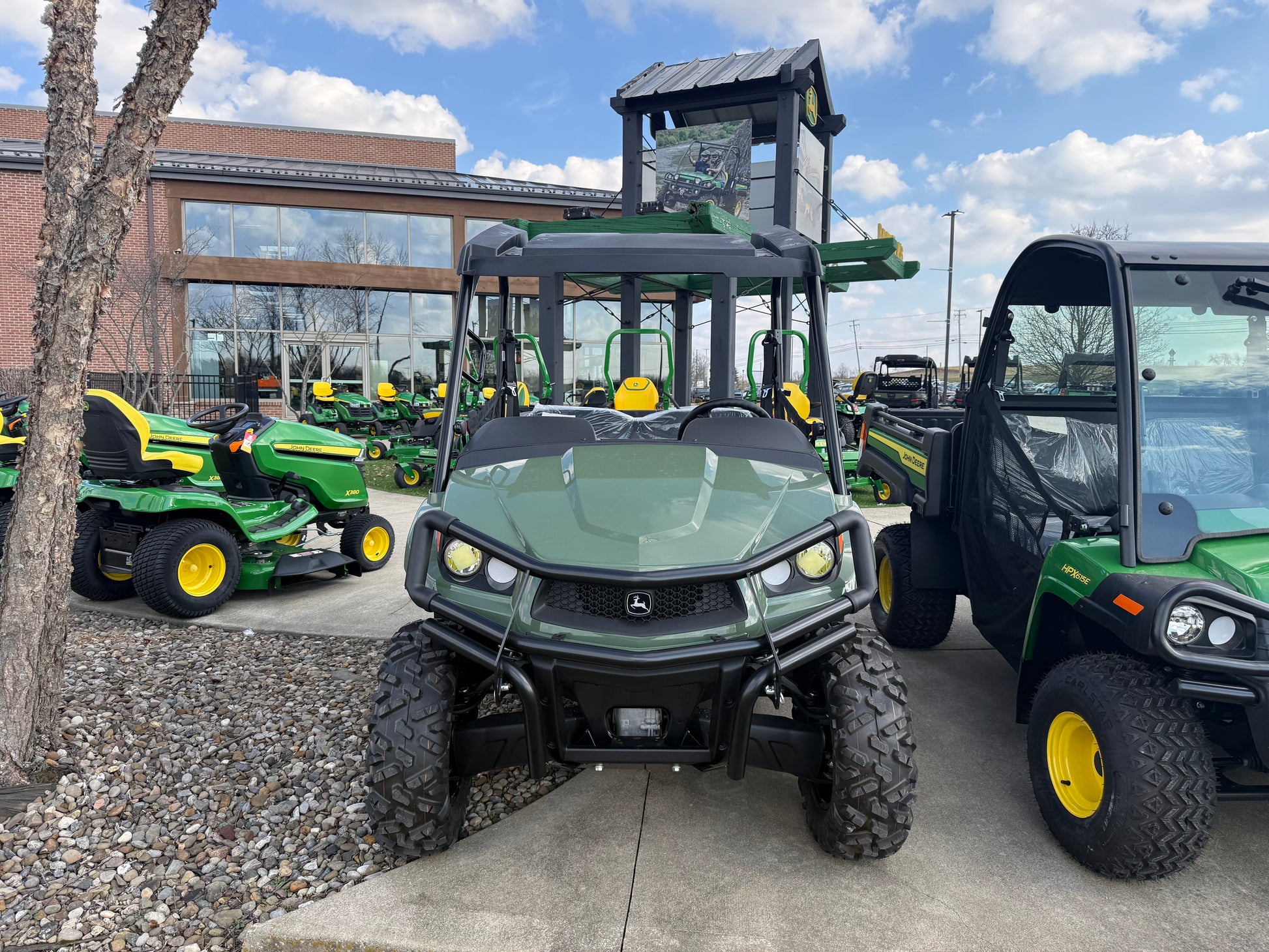 Front view of a green mid-size John Deere crossover utility vehicle with rugged tires and protective roof displayed outdoors on concrete near other John Deere equipment.