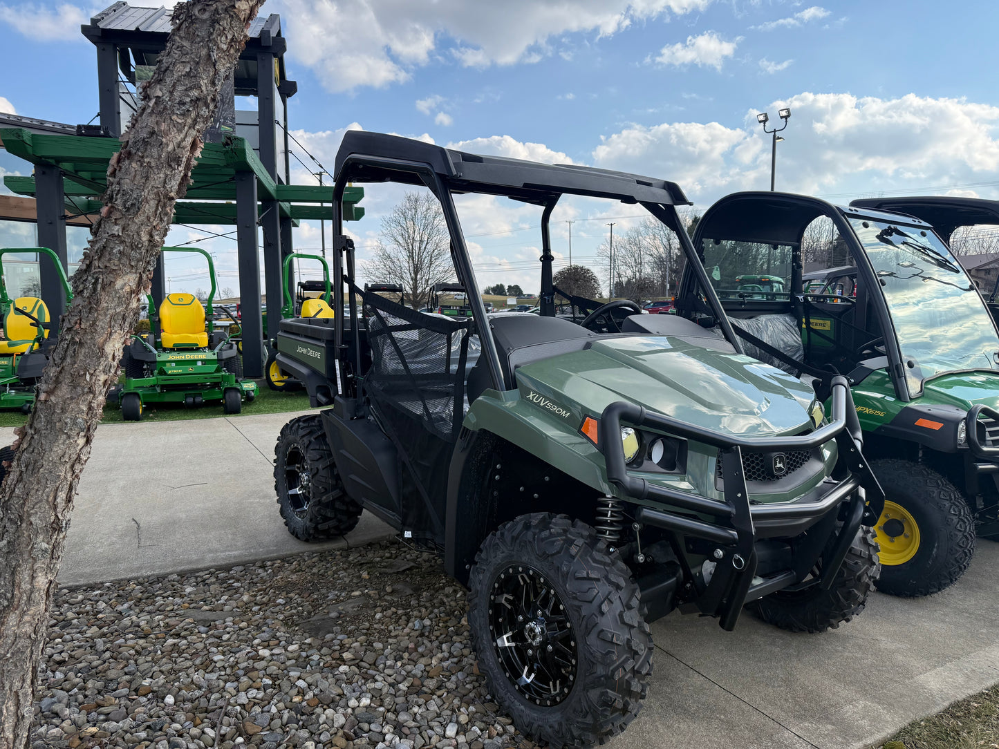 Green mid-size John Deere utility vehicle with aggressive off-road tires and protective roof parked outdoors near other equipment at dealership lot.