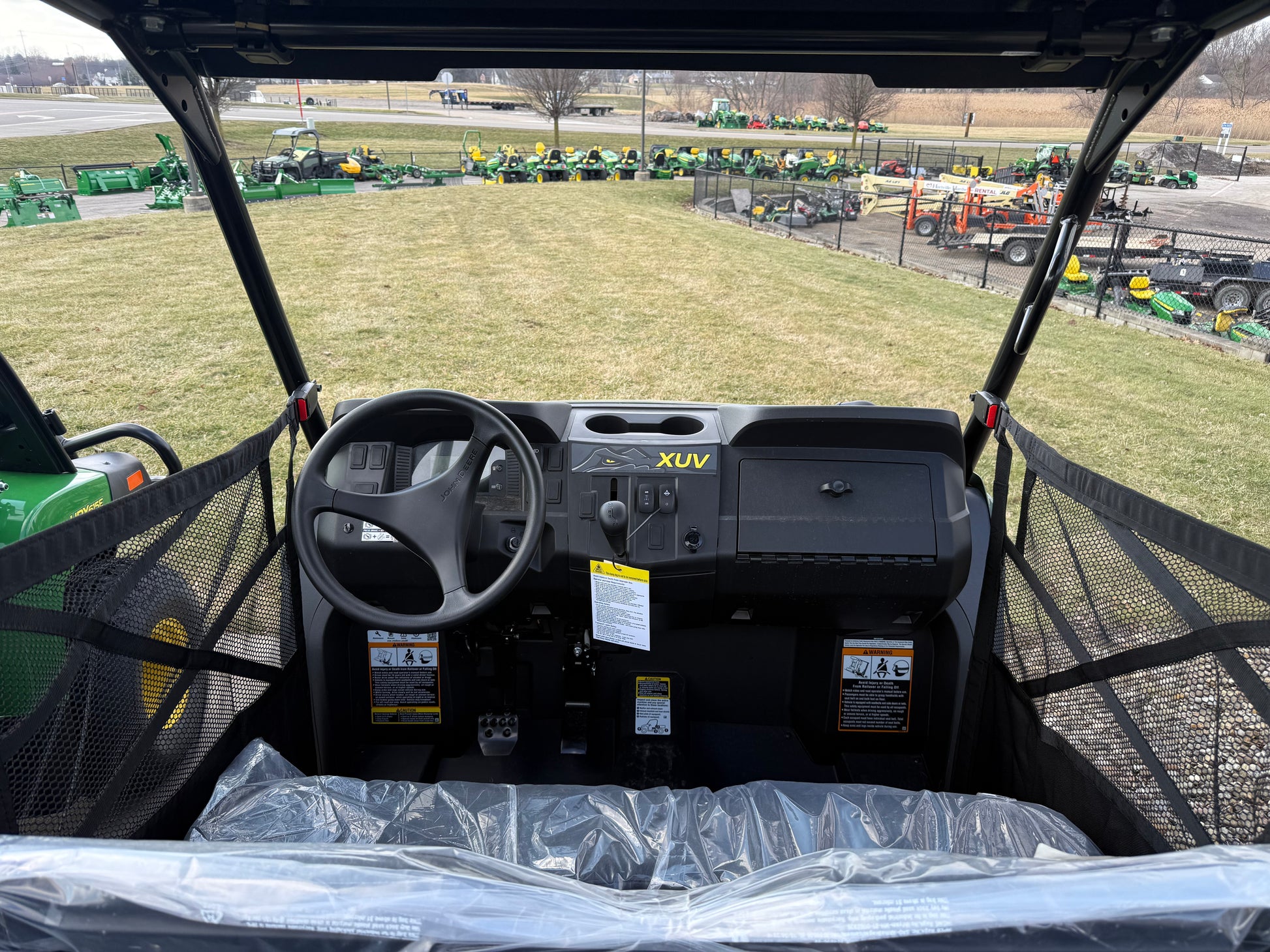 Interior view of a John Deere mid-size crossover utility vehicle showing steering wheel, dashboard controls, and plastic-covered seating in an outdoor dealership setting.