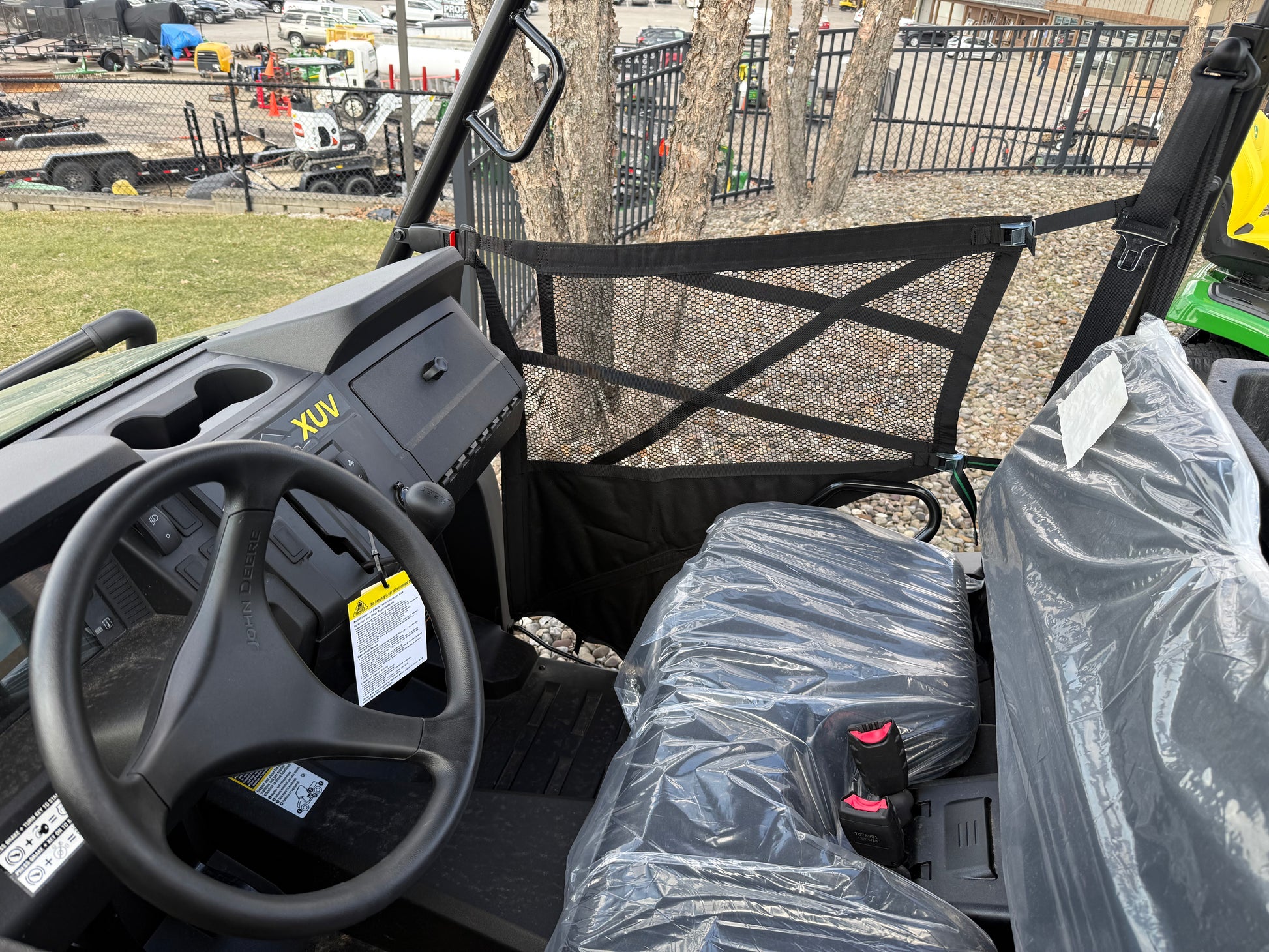 Interior view of a John Deere mid-size crossover utility vehicle showing steering wheel, dashboard, and plastic-covered seats with mesh side door in an outdoor lot.