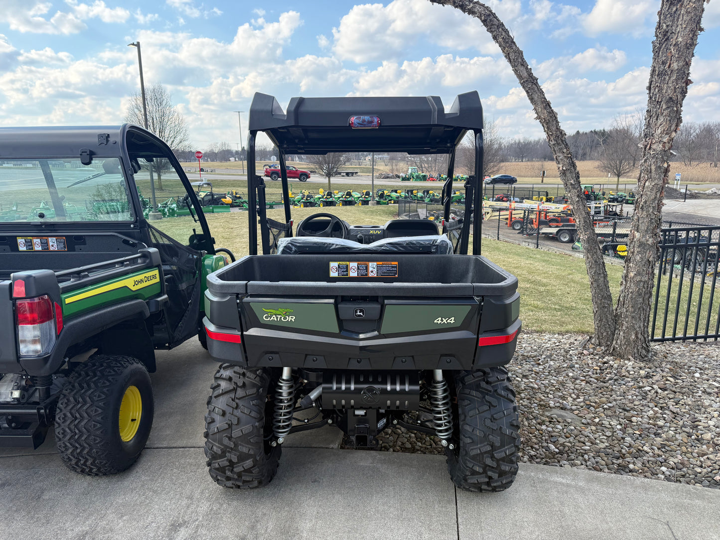 Rear view of a green John Deere mid-size crossover utility vehicle featuring rugged tires, suspension, and cargo bed parked outdoors near other equipment at dealership lot