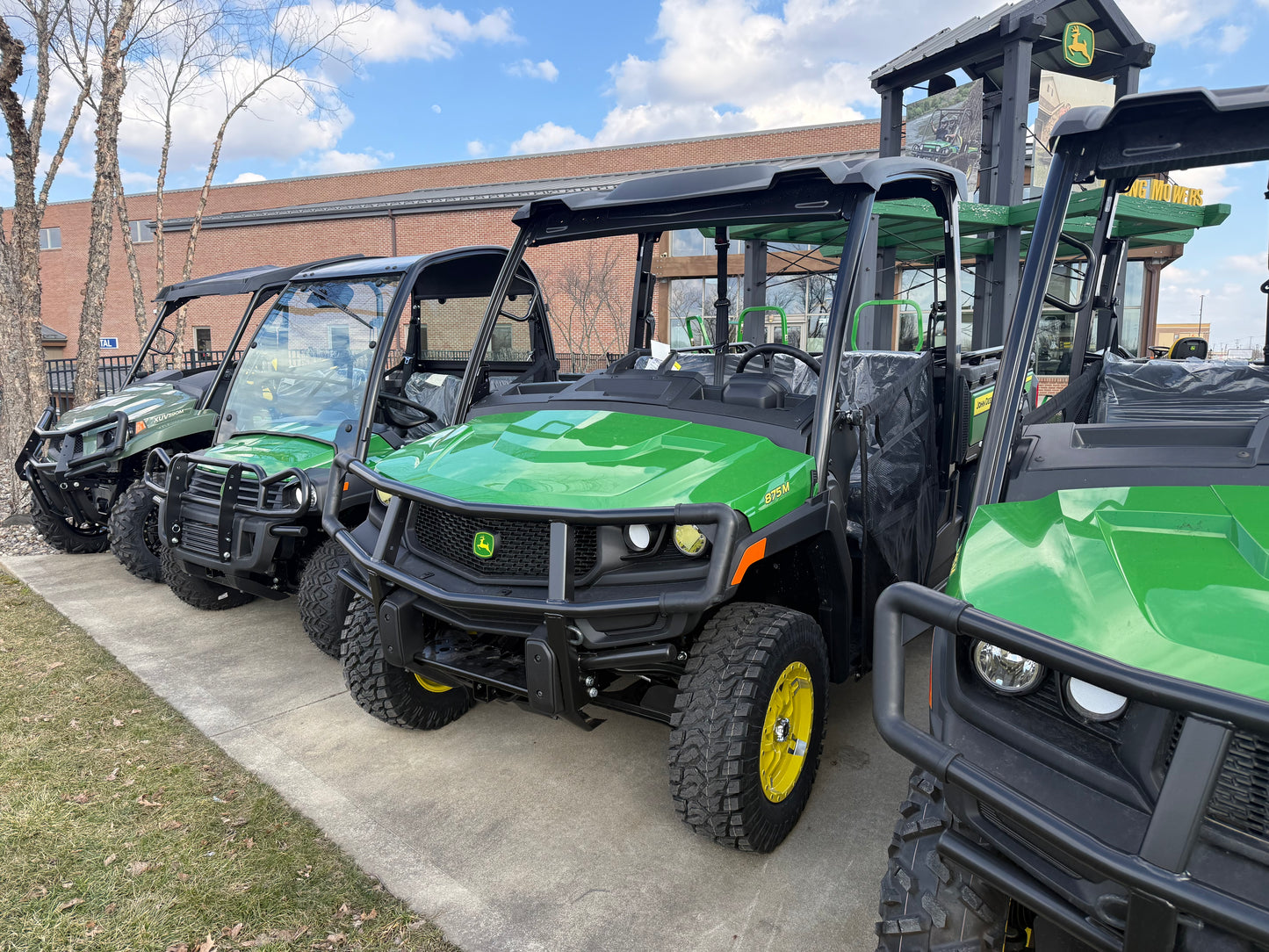 Row of green John Deere diesel crossover utility vehicles parked outdoors on concrete near a grassy strip under a partly cloudy sky at dealership lot