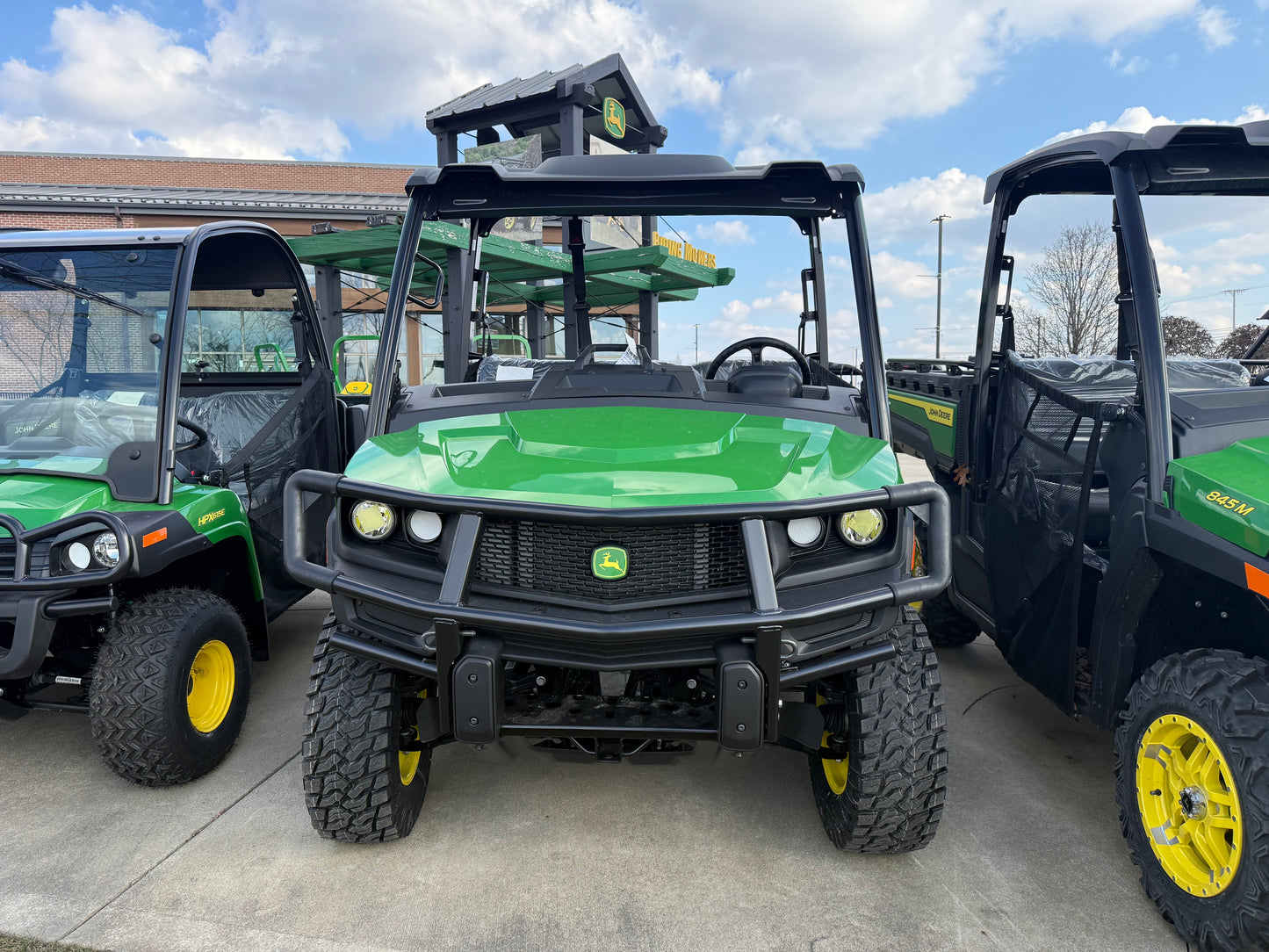 Front view of a green John Deere diesel crossover utility vehicle with heavy-duty grille and rugged tires parked among other John Deere UTVs at dealership lot under partly cloudy sky