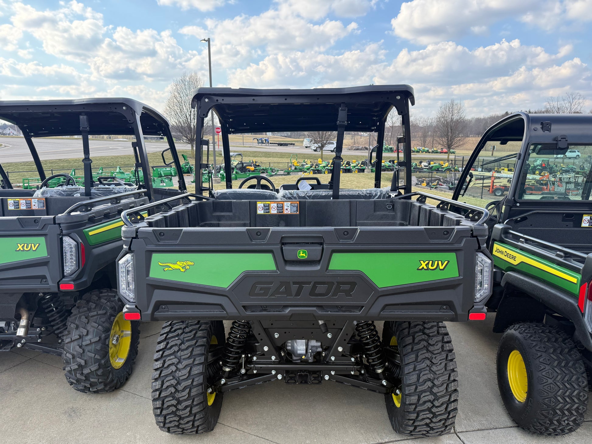 Rear view of a green John Deere Gator XUV utility vehicle with rugged tires and spacious cargo bed, parked outdoors with other UTVs in display lot