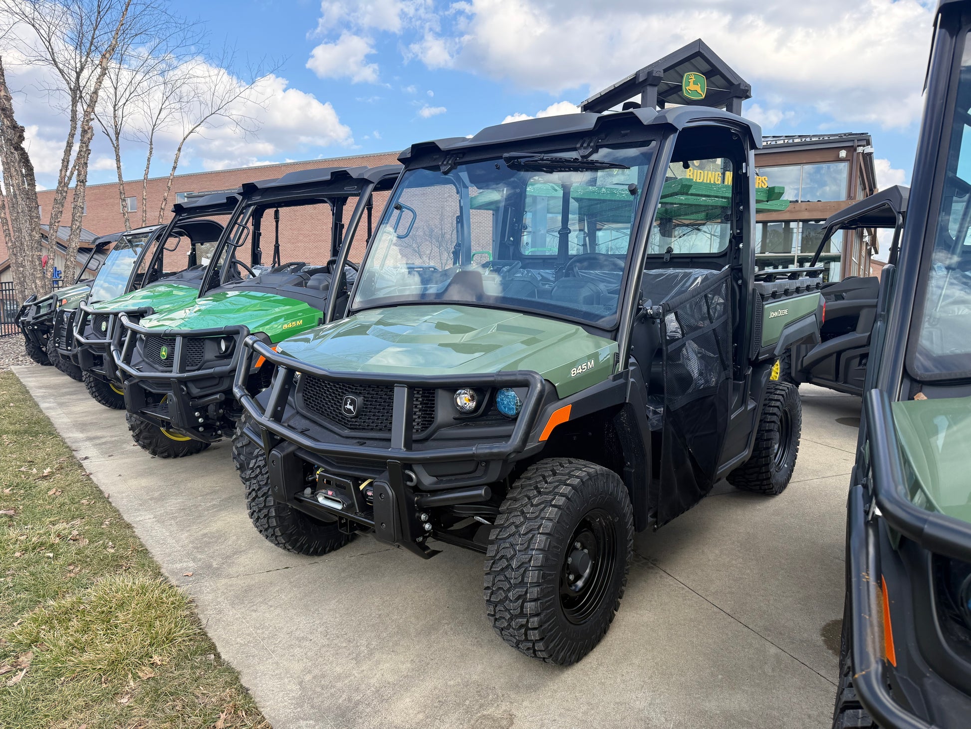 Line of green John Deere utility vehicles parked outdoors on concrete pavement near grassy area under blue sky with scattered clouds, in a John Deere UTVs collection display.