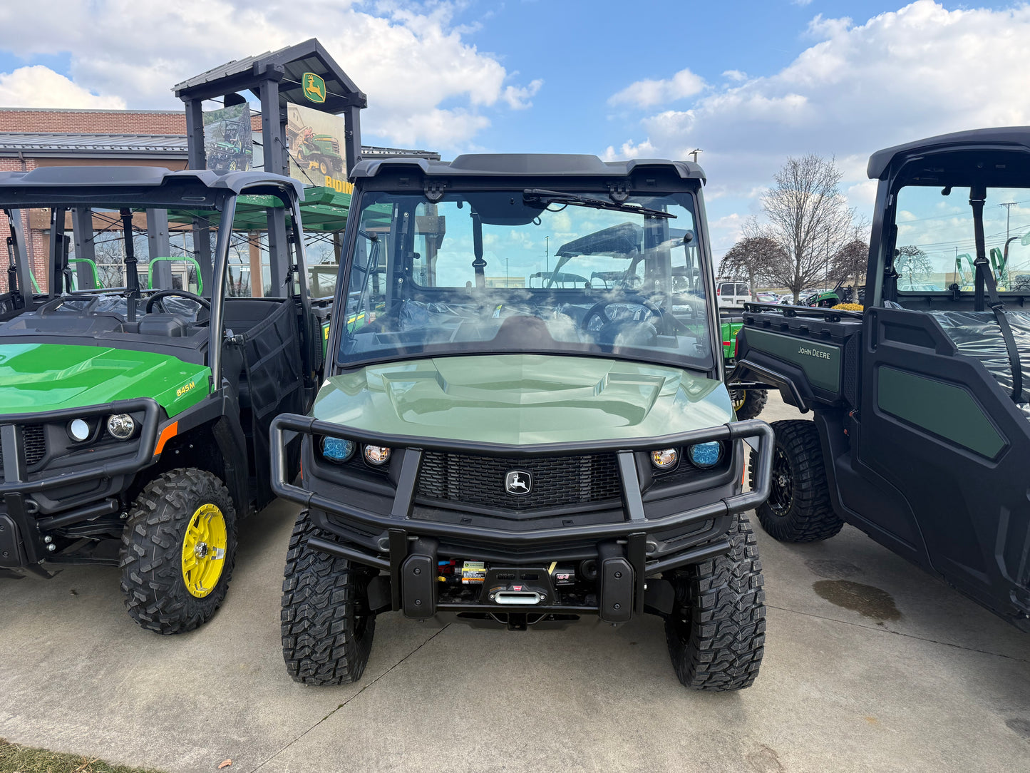 Front view of a green John Deere crossover utility vehicle with protective grille and rugged tires parked outdoors among other John Deere UTVs and equipment at dealership.