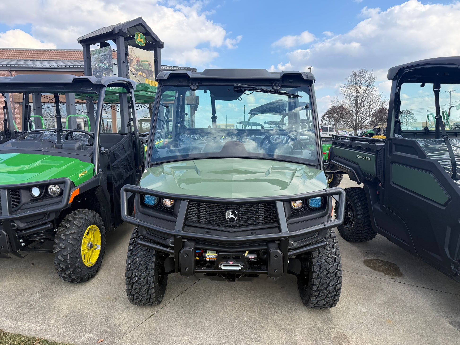 Front view of a green John Deere crossover utility vehicle with protective grille and rugged tires parked outdoors among other John Deere UTVs and equipment at dealership.