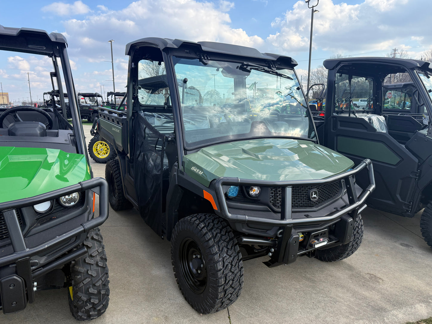 Three John Deere utility vehicles parked outdoors on concrete pavement with a partly cloudy sky, showcasing rugged tires and protective frames.