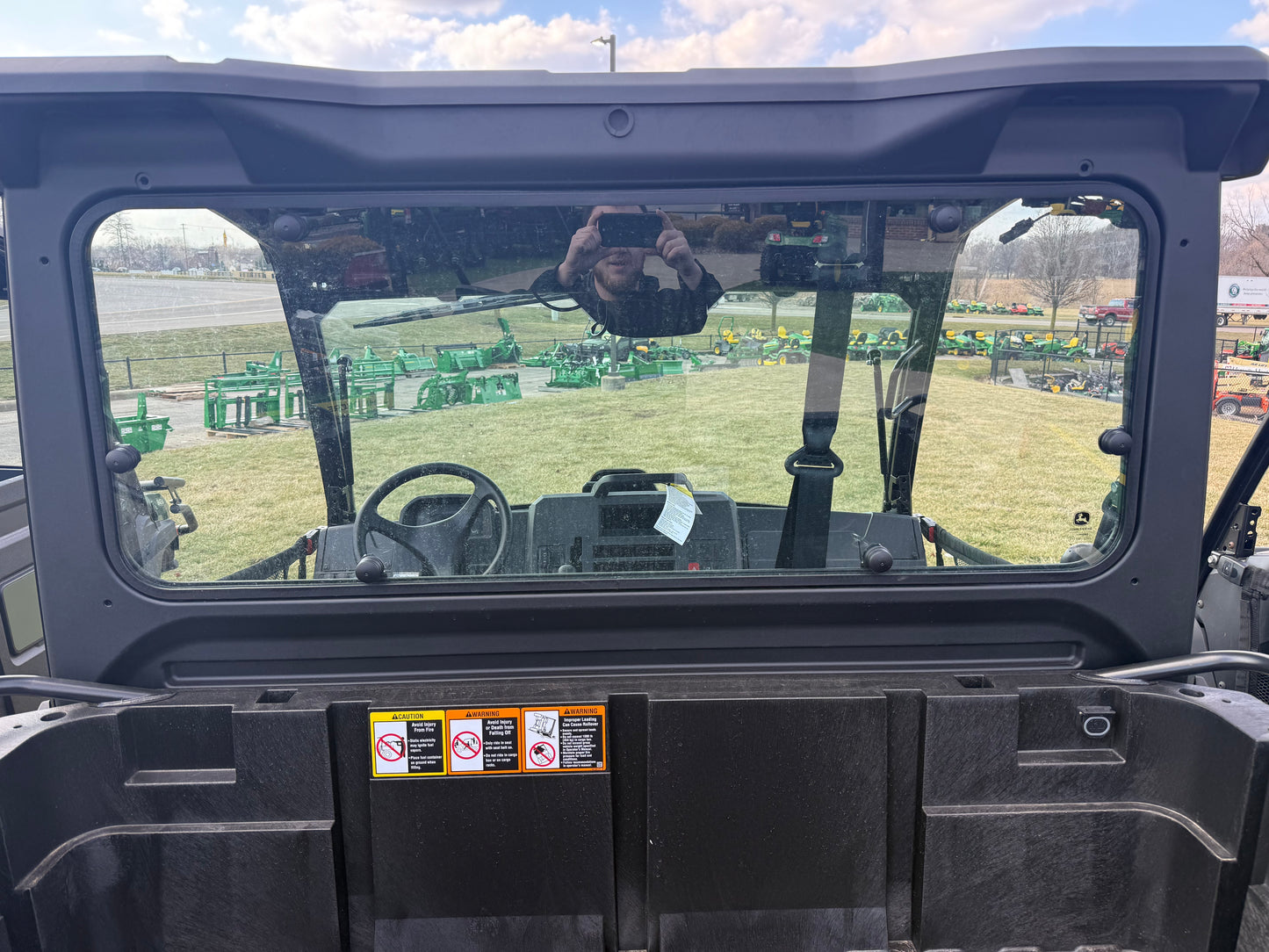 View through the rear window of a John Deere utility vehicle showing the enclosed cab interior and steering wheel in an outdoor equipment lot display.