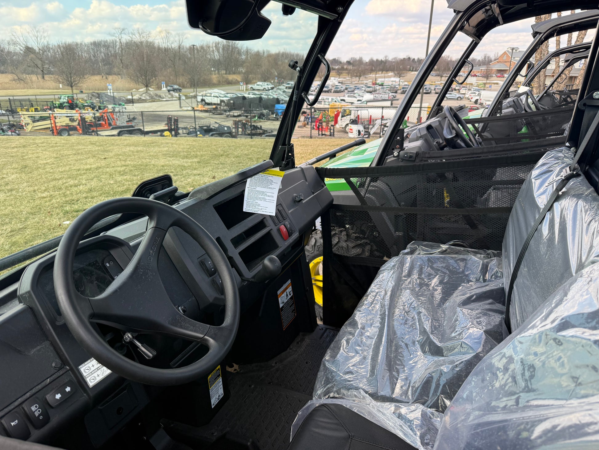 Interior view of John Deere utility vehicle showcasing steering wheel, dashboard, and protective plastic-covered seating at outdoor dealership lot with equipment in background