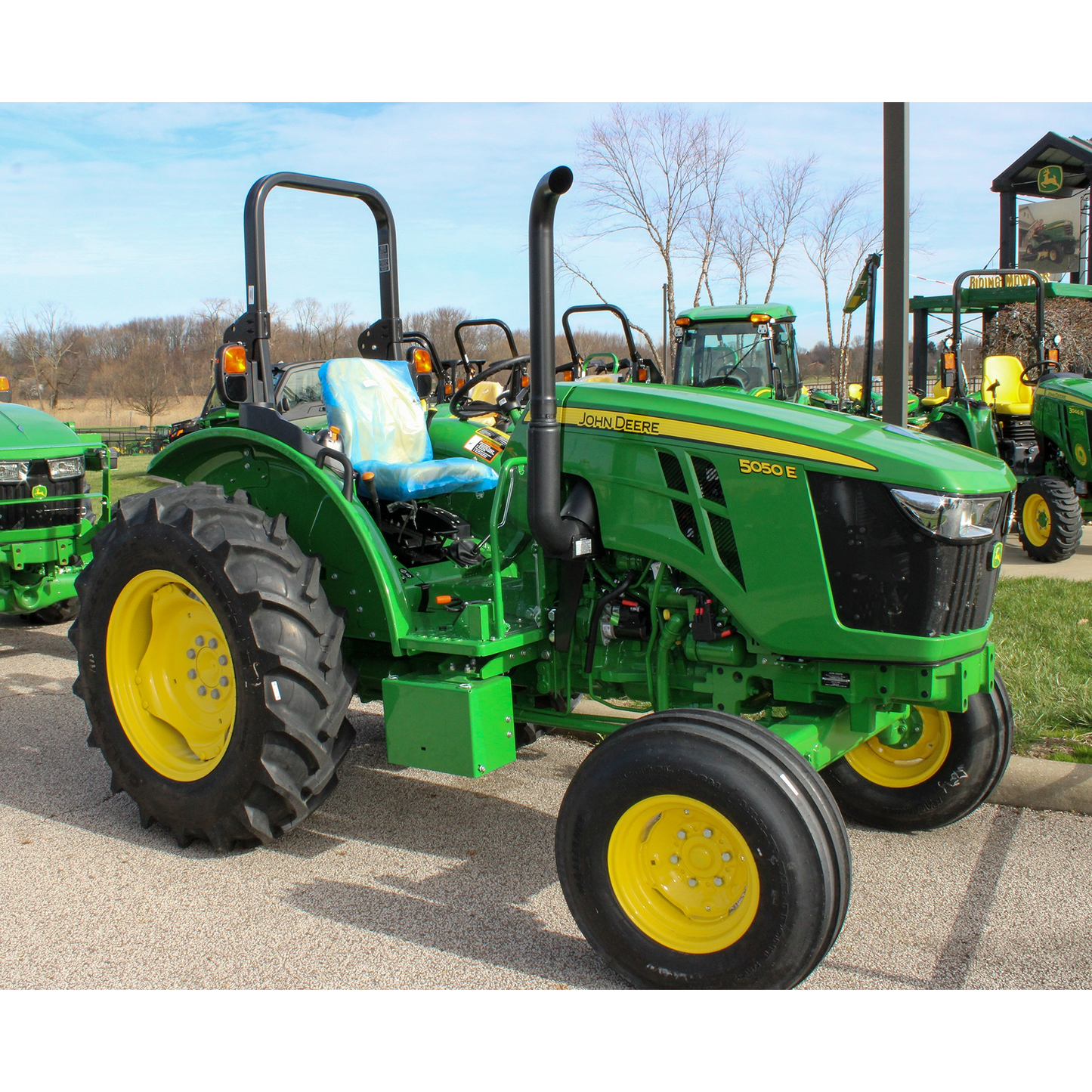 Green and yellow compact utility tractor with large rear tires and operator seat, displayed outdoors with other John Deere equipment at Hartville Hardware in the lawn and garden section