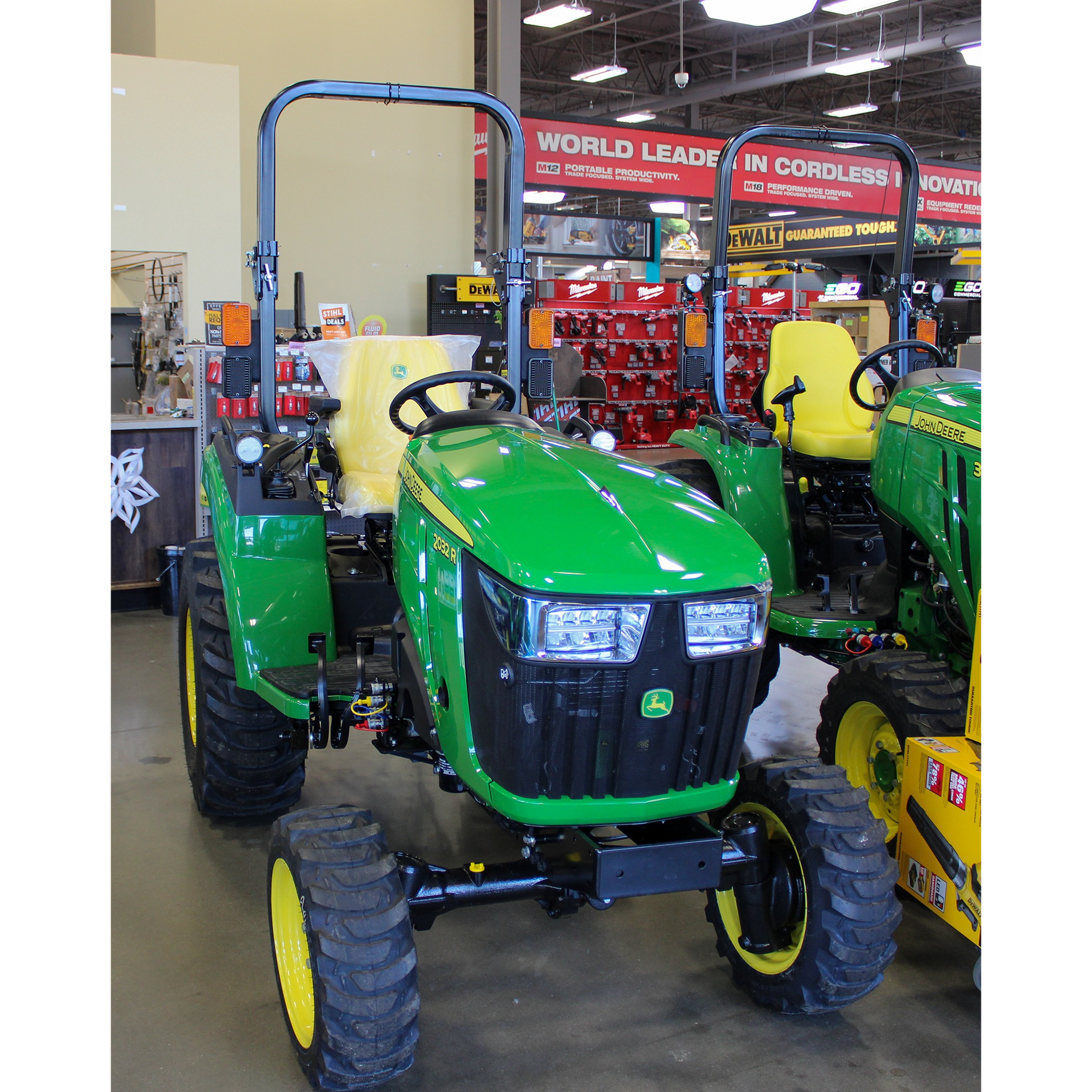 Green compact John Deere tractor with yellow wheels and rollover protection, displayed indoors among outdoor power equipment and tools collections.