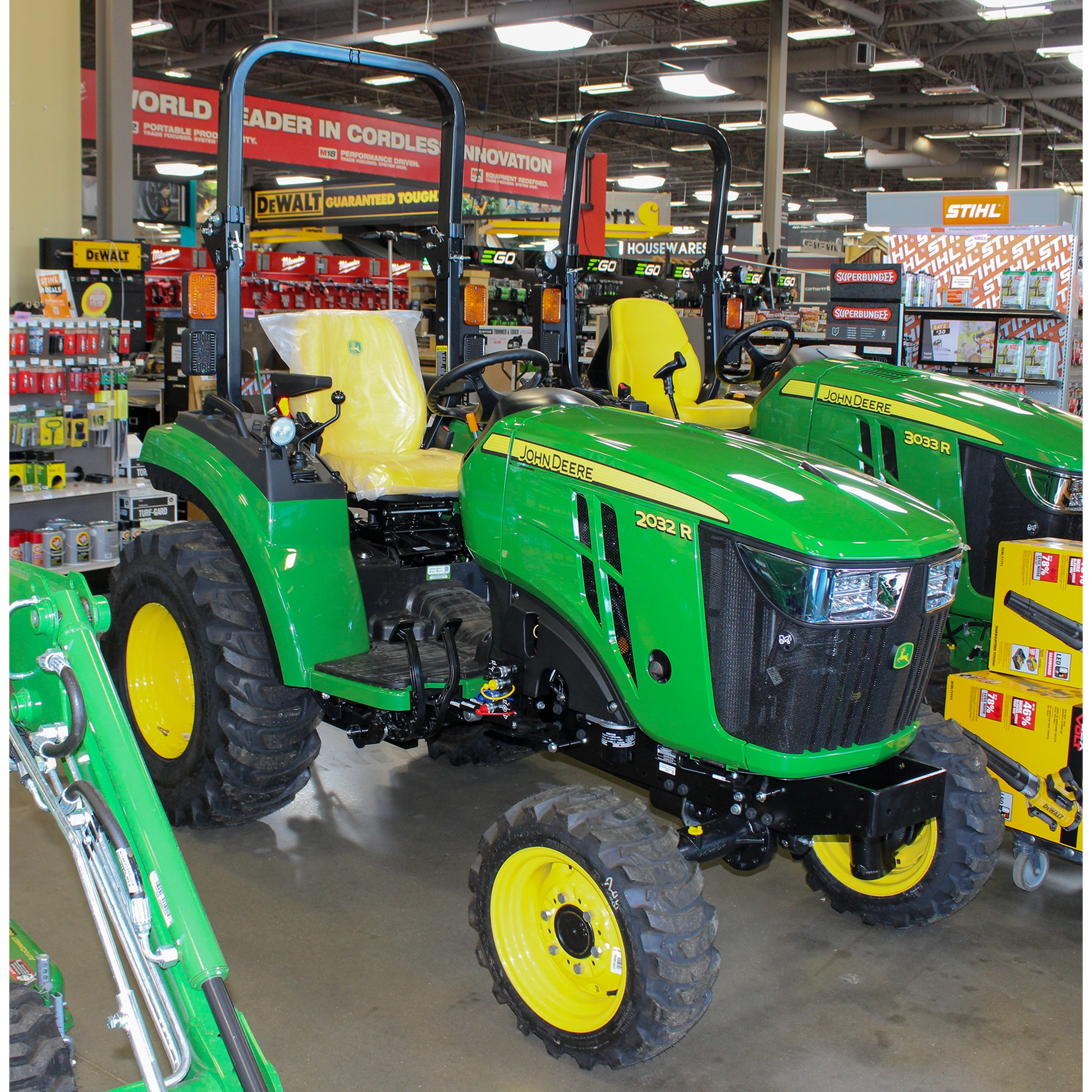 Green John Deere 2032 R compact utility tractor with yellow wheels, rollover protection, and rugged tires displayed indoors among outdoor power equipment collections.