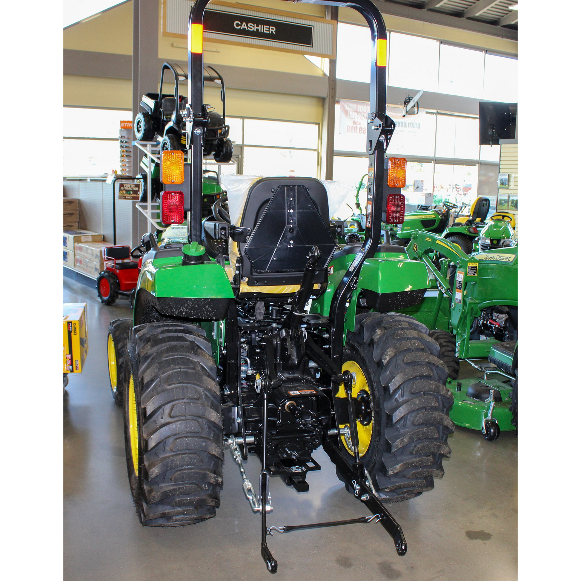 Rear view of a green John Deere compact utility tractor with yellow wheels, heavy tread tires, and rear hitch, displayed indoors in an outdoor power equipment collection.