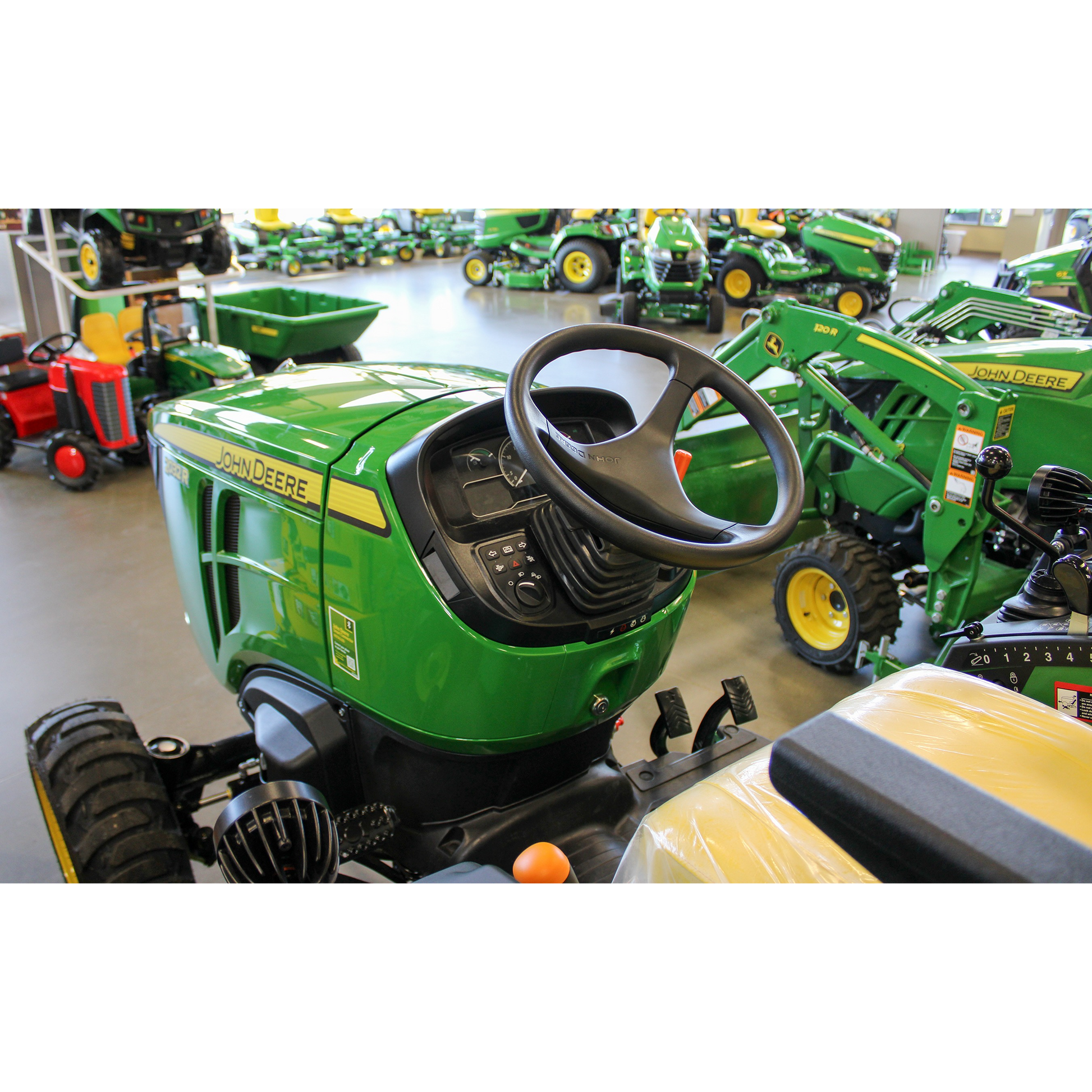 Green John Deere compact utility tractor featuring a detailed dashboard, steering wheel, and yellow seat, displayed in an indoor equipment showroom among outdoor power tools and lawn & garden machinery.