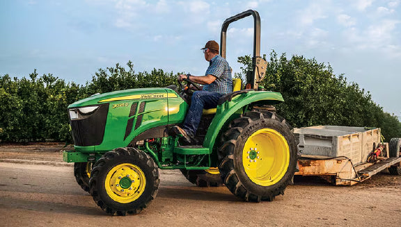 Man driving a green and yellow tractor pulling a trailer on a dirt path with trees and a partly cloudy sky in the background