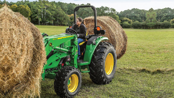 Person operating a green tractor with yellow wheels lifting a large round hay bale in a grassy field bordered by trees under a cloudy sky
