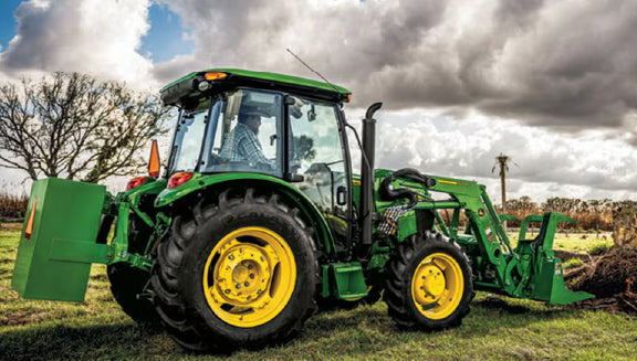 Green tractor with yellow wheels parked on grass under a dramatic cloudy sky with bare trees in the background
