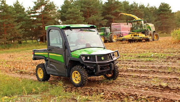 Green utility vehicle with enclosed cab parked on a field with trees and farming machinery harvesting crops in the background