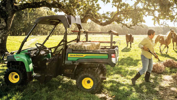 Green utility vehicle with yellow wheels parked on a sunlit grassy field while a person spreads hay near horses grazing in the background